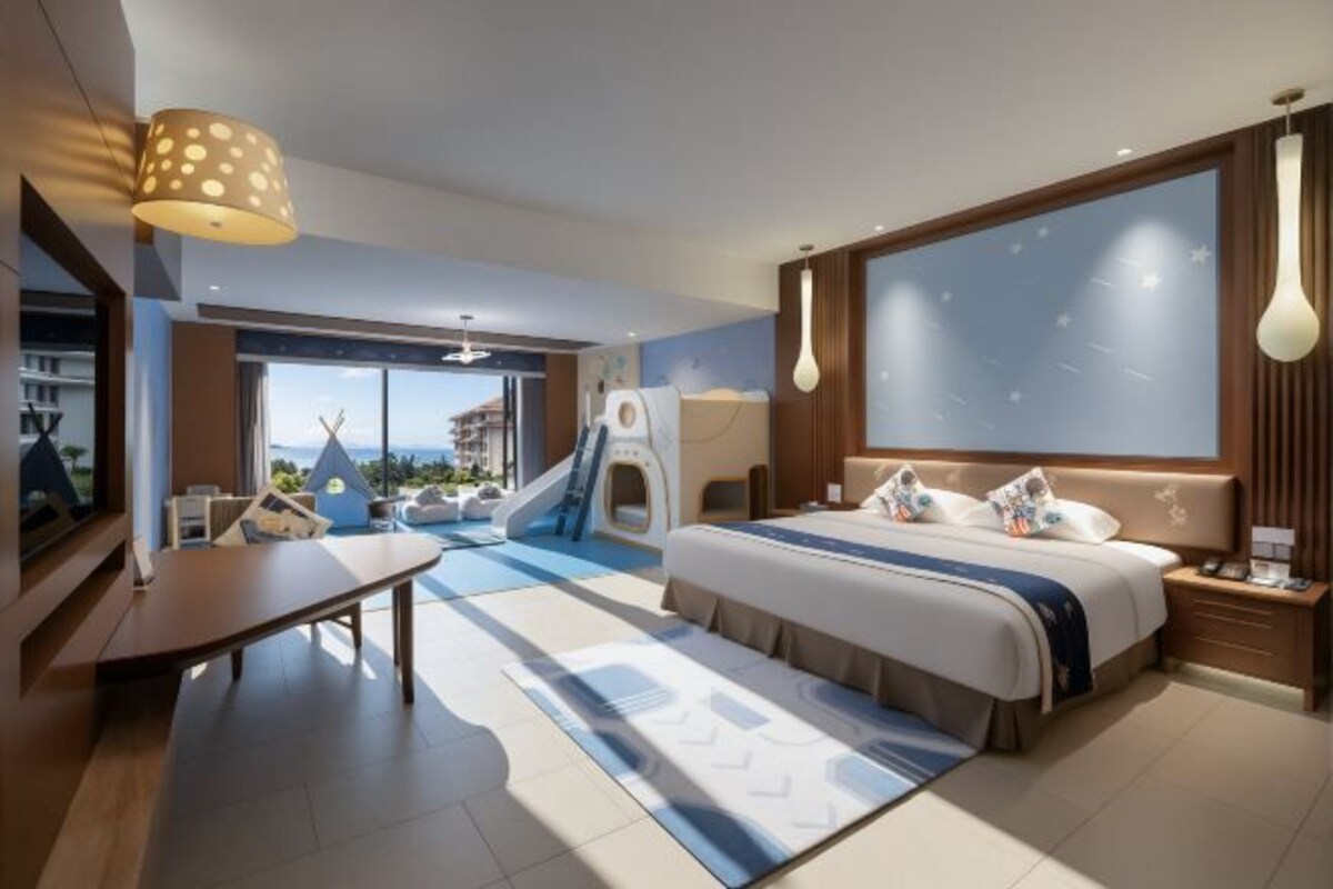 A modern hotel room with a bed, bedside table, lamp, and a view of a slide and pool.