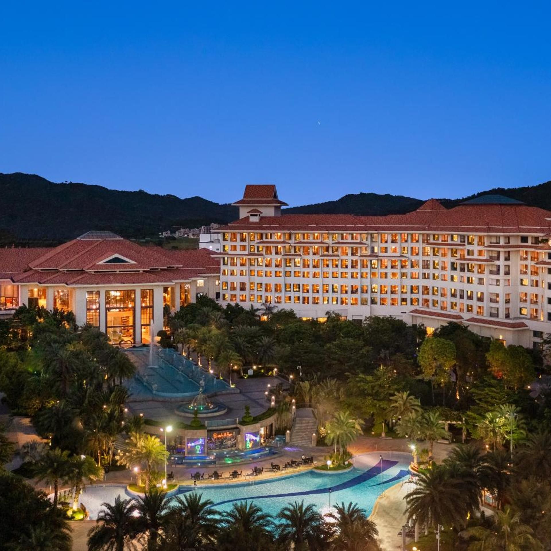 A large hotel with multiple floors, a swimming pool, and a landscaped area with palm trees.