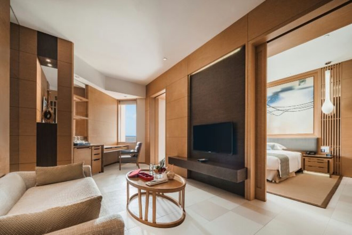 A modern hotel room with a large TV, a sofa, a dining table, and a separate bedroom area.