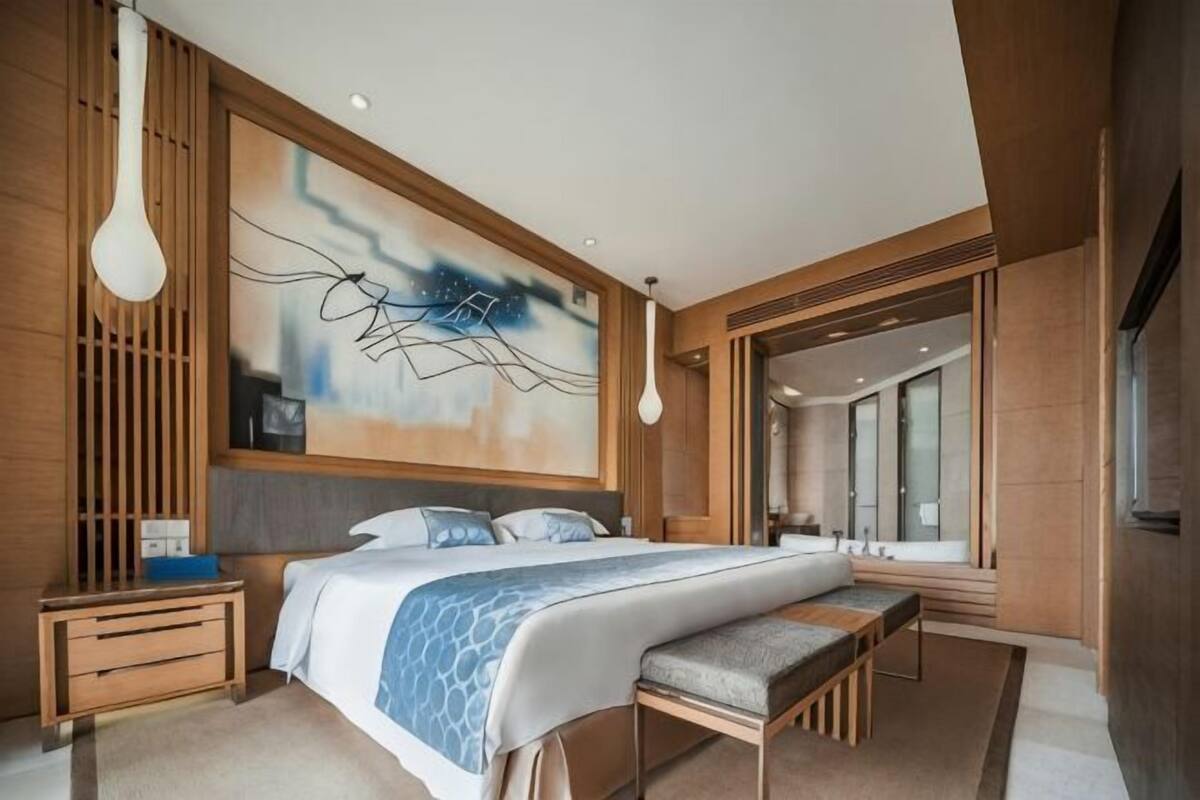 A modern hotel room with a large bed, wooden paneling, a bench, a nightstand, and a large abstract painting.