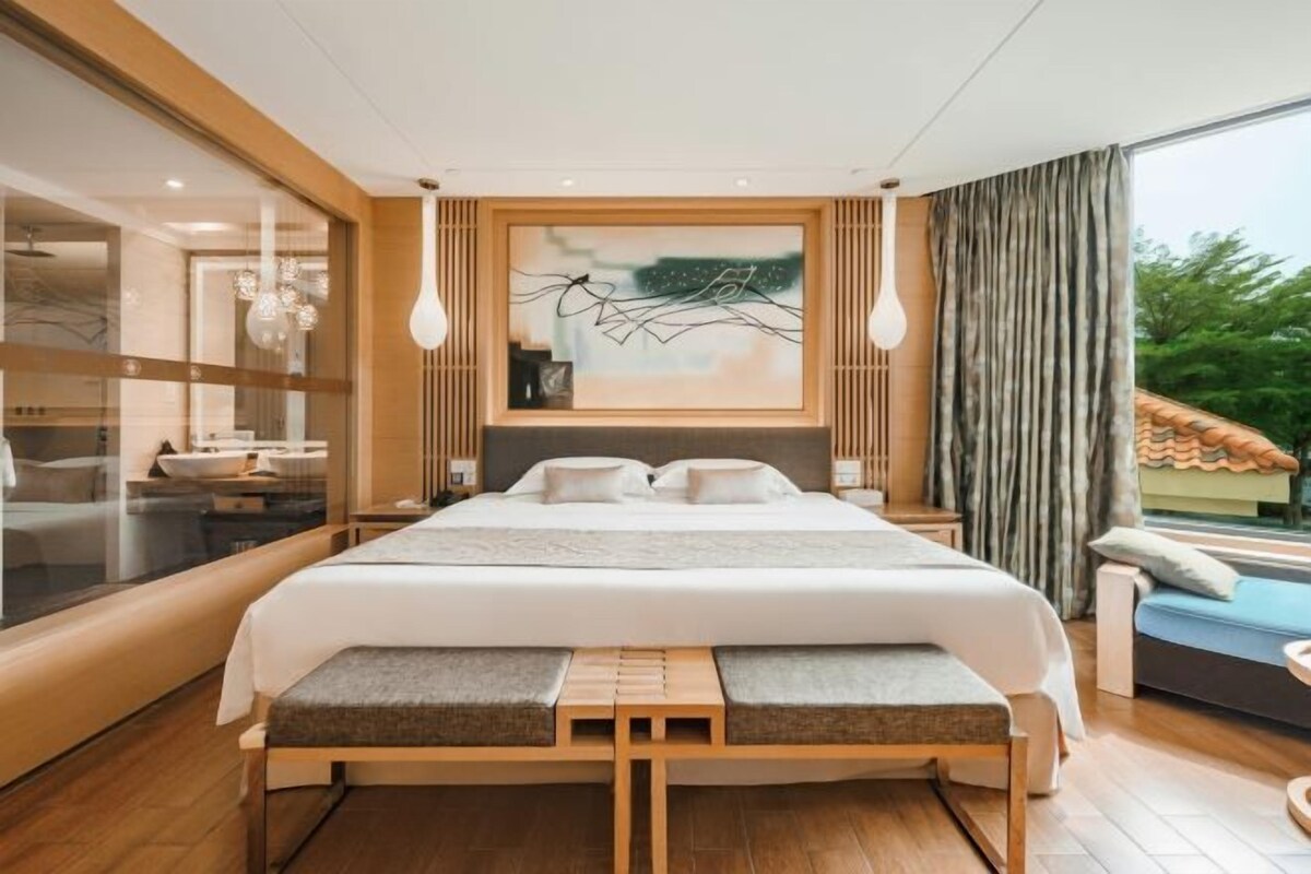 A modern bedroom with a large bed, a bench, a painting, and a view of the outdoors.