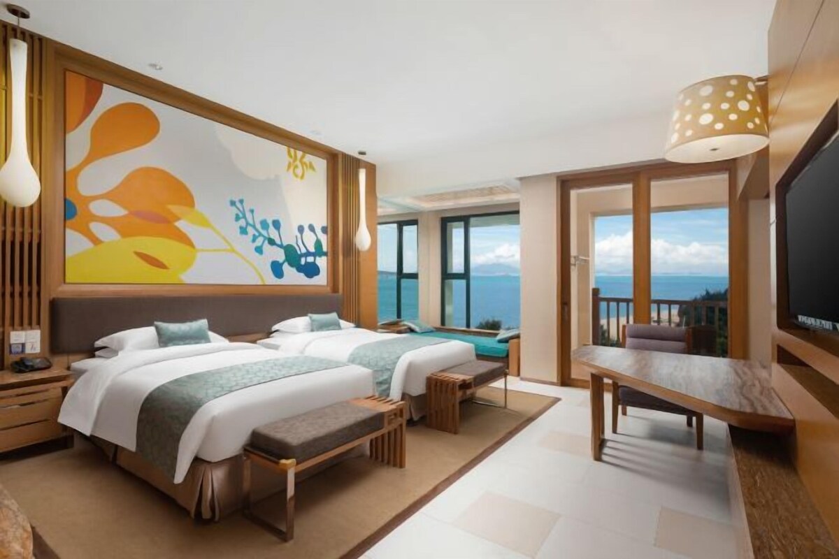 A modern hotel room with two beds, a dining area, and a large window offering a view of the sea.