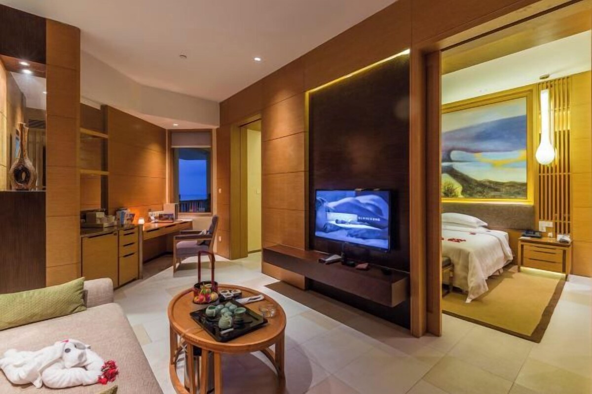 A modern hotel room with a TV, sofa, dining area, and a painting of a landscape.