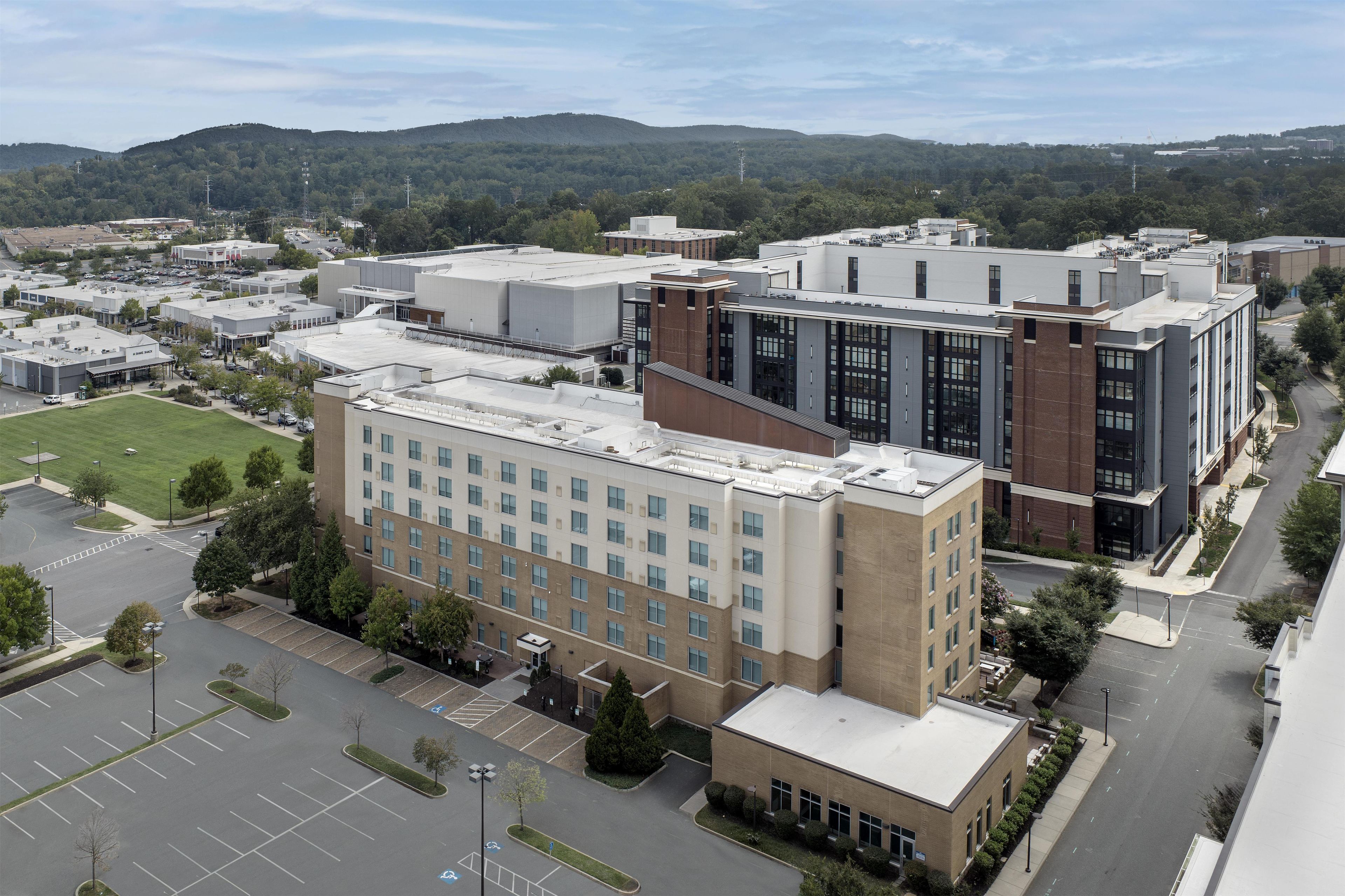 A multi-story building with a flat roof, surrounded by a parking lot and a green space.