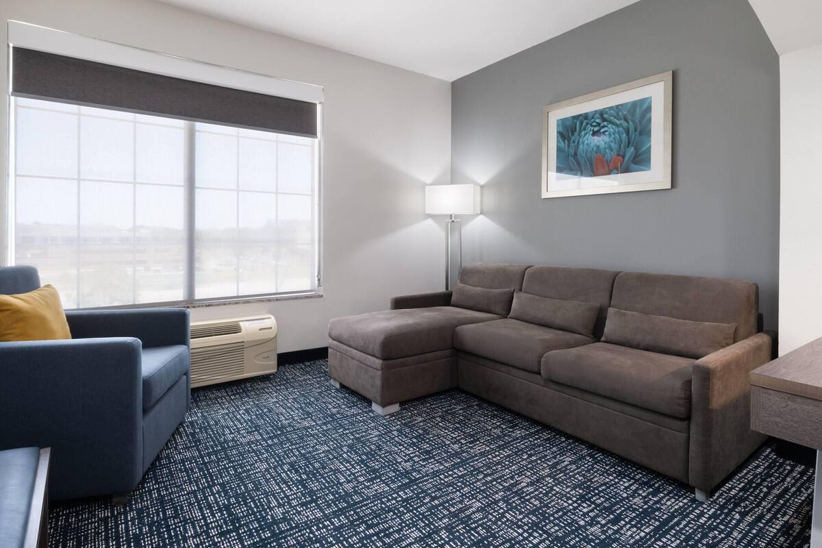 Une chambre d’hôtel moderne, dotée d’un canapé d’angle, d’un petit fauteuil, d’un lampadaire et d’une grande fenêtre donnant sur la ville.