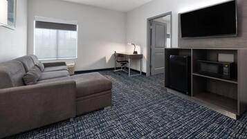A hotel room with a gray sofa, a desk with a chair, a flat-screen TV, a microwave, and a refrigerator.
