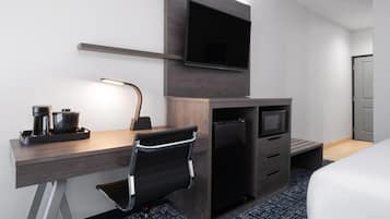 Habitación de hotel con escritorio de madera, una silla de oficina negra, televisor de pantalla plana montado en la pared, mini refrigerador y cama con ropa blanca.