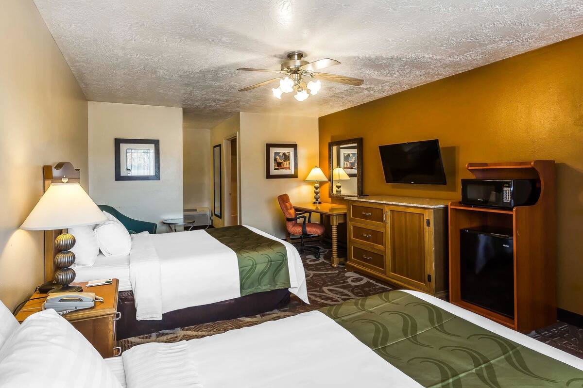 A hotel room with two beds, a desk with a chair, a television, a microwave, and a ceiling fan.