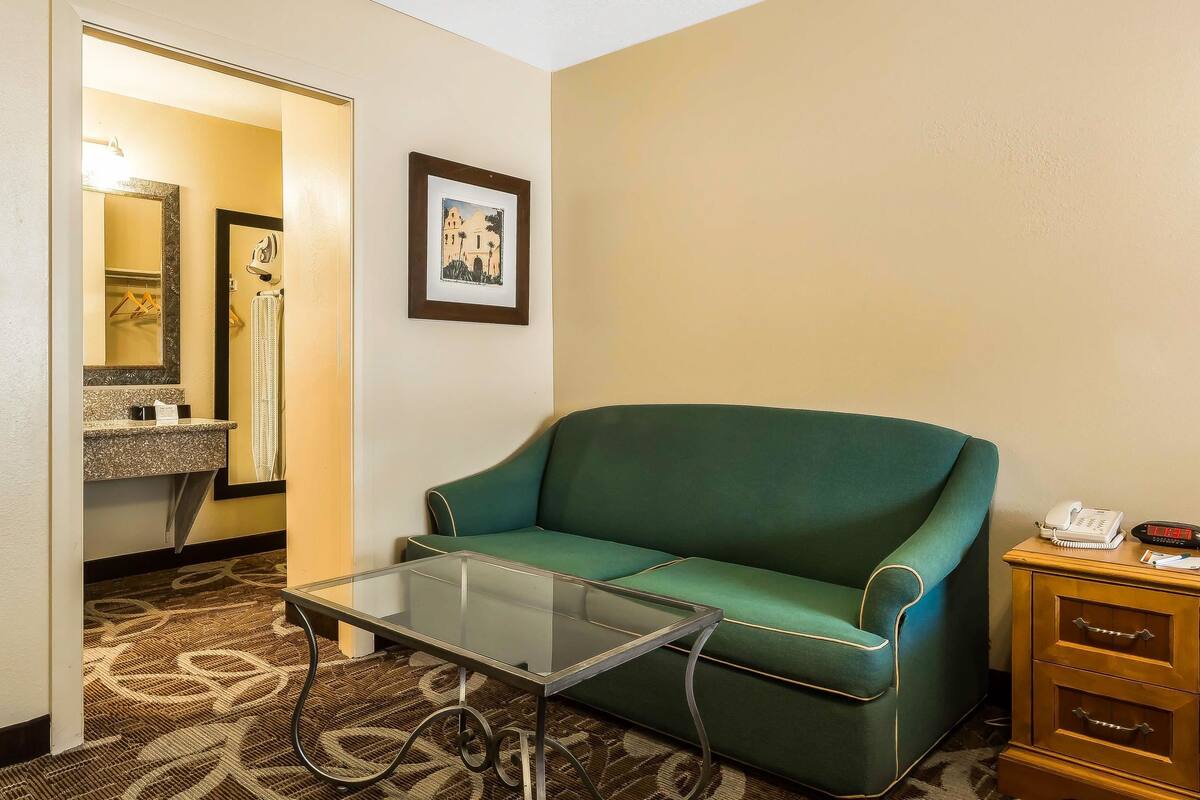 A hotel room with a green sofa, a glass coffee table, a wooden nightstand with a phone, a framed picture on the wall, and a bathroom with a sink and mirror.