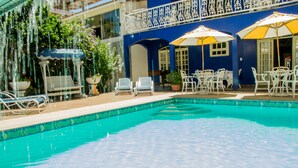 Une piscine entourée de chaises longues et de parasols, le tout bordé d’un bâtiment bleu aux balustrades blanches.