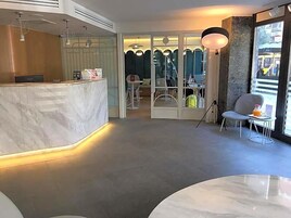 A modern hotel lobby with a marble reception desk, a seating area with a table and chairs, and large windows.