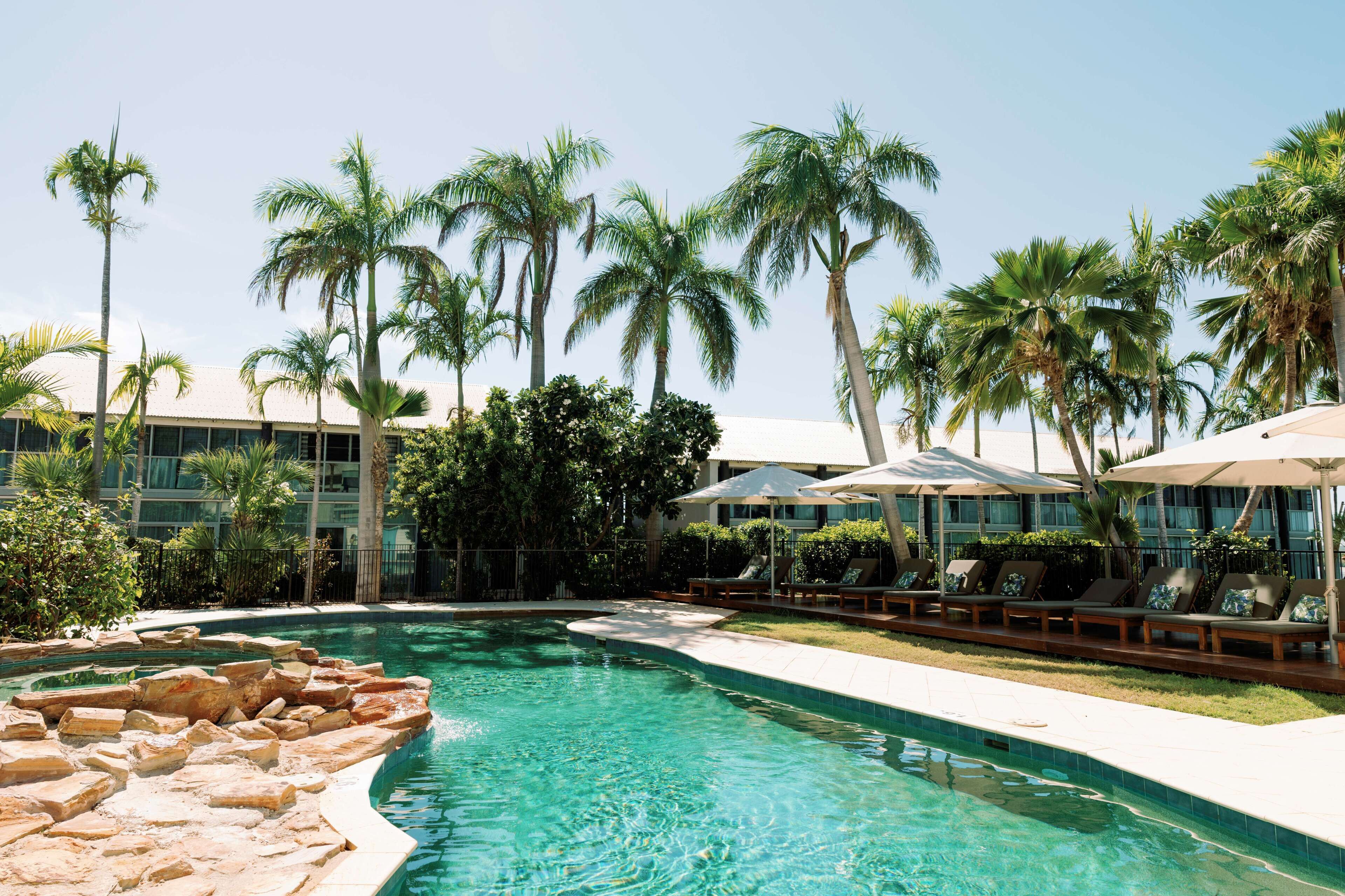 A swimming pool with lounge chairs and umbrellas, surrounded by palm trees and a building.