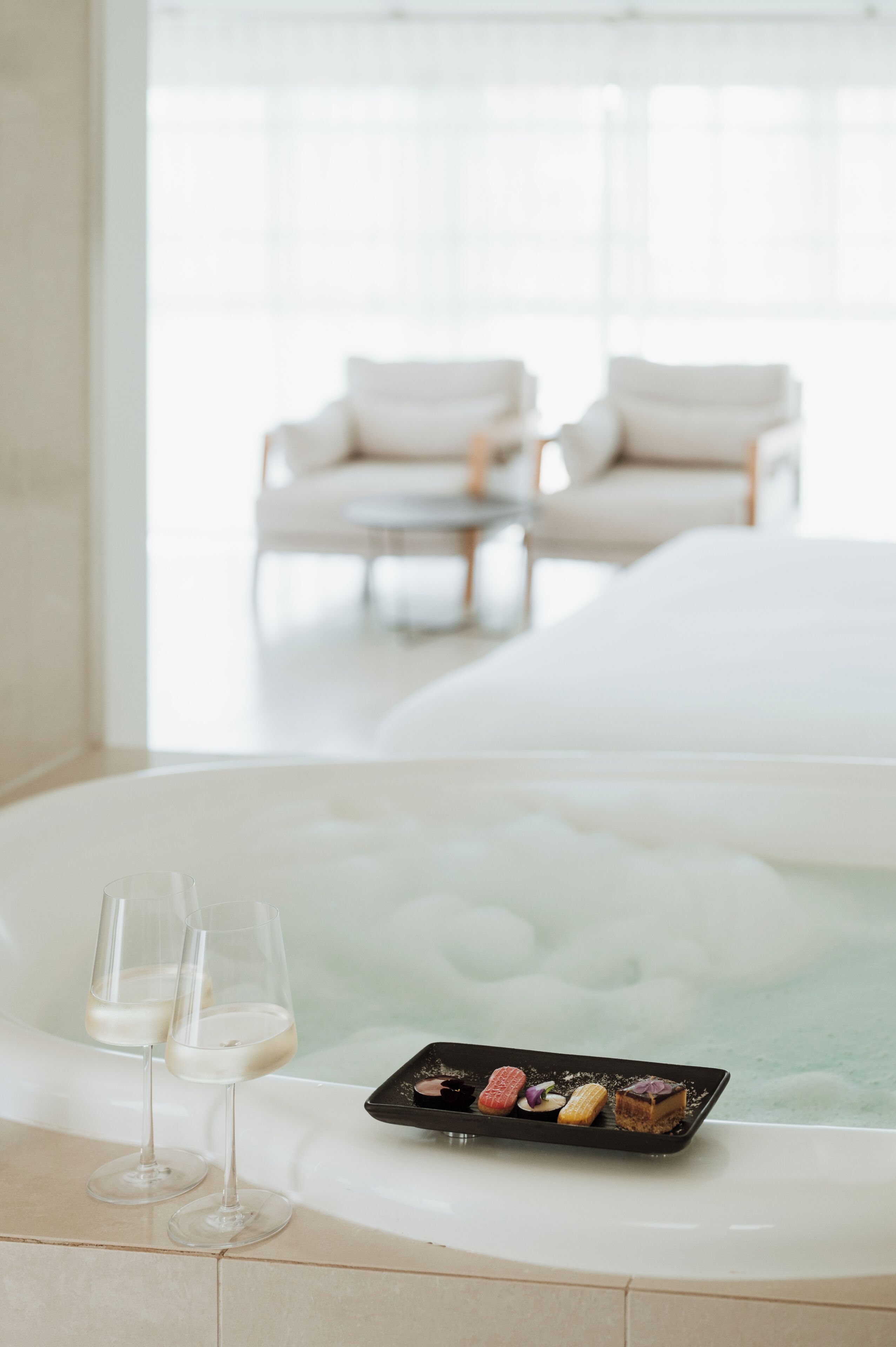 A spa tub with two glasses of white liquid, a tray of assorted desserts, and a view of a white room with a chair and a table.