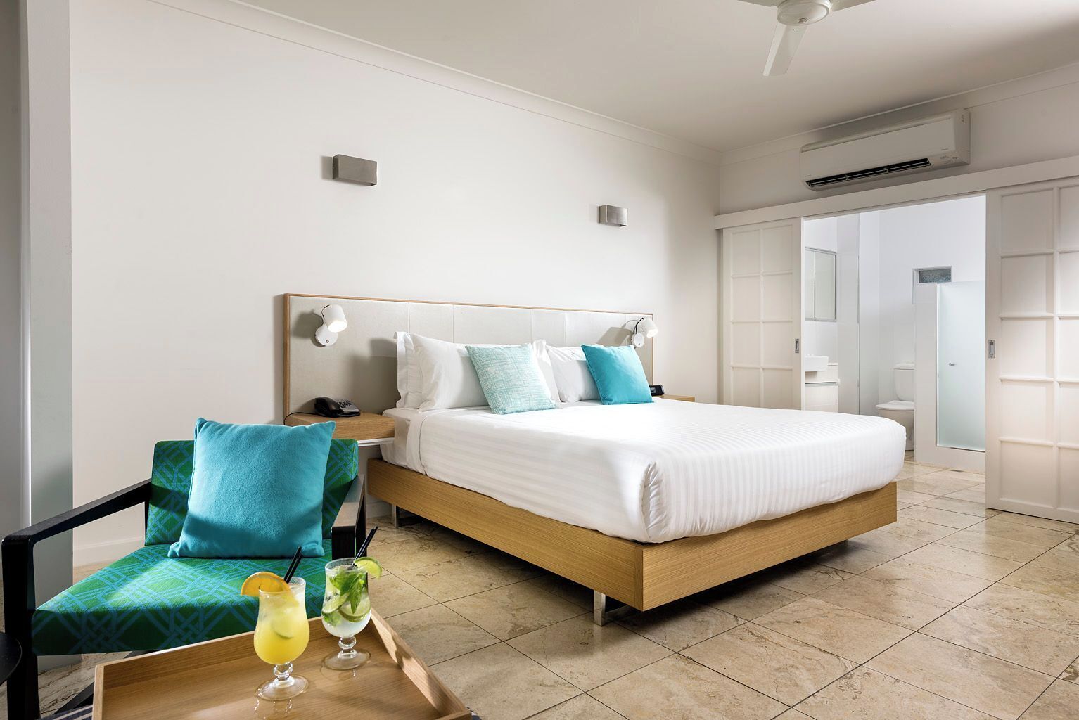 A modern hotel room with a large bed, a bedside table with a phone, a chair with a patterned cushion, and a tray with drinks.