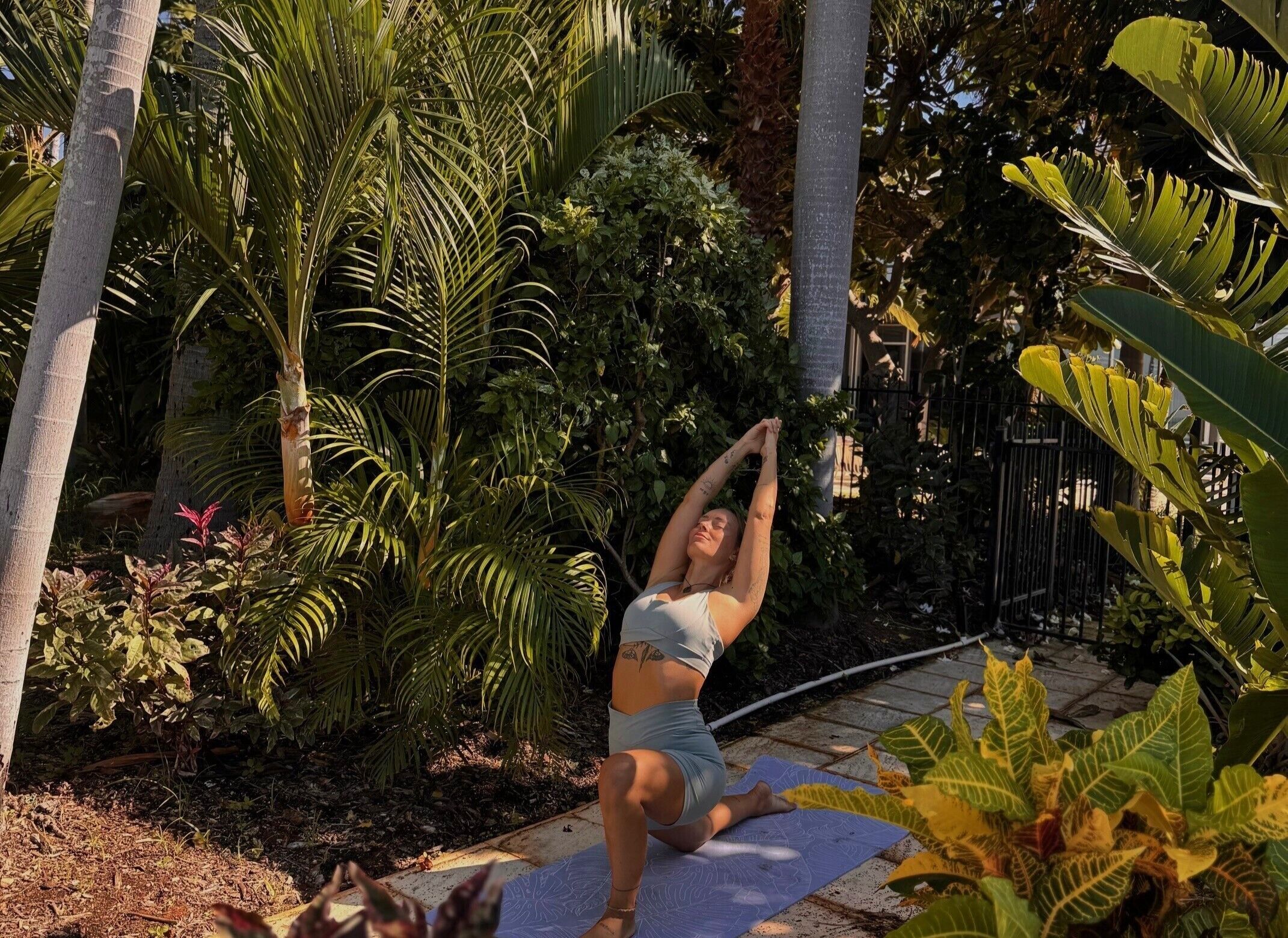 Una persona realizando una postura de yoga sobre una esterilla en un jardín con diversas plantas y árboles.