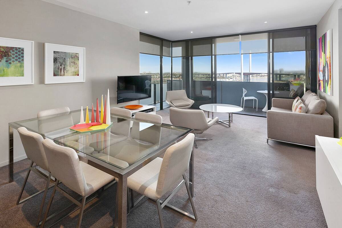 A modern living room with a glass dining table, comfortable seating, a flat-screen TV, and large windows offering a city view.