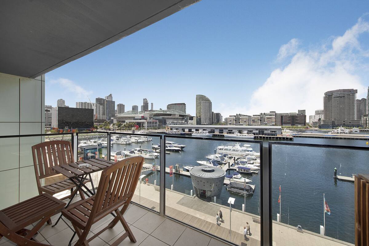 A balcony with wooden chairs and a table overlooking a marina with boats and a city skyline.