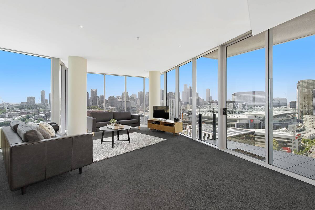 A modern living room with a large sofa, a coffee table, and a TV on a wooden stand. The room has large windows offering a city view.