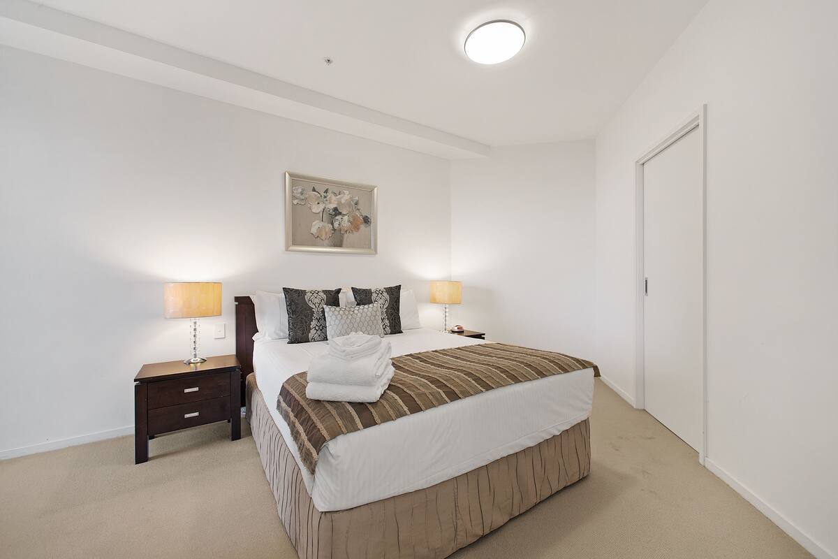 A neatly made bed with a striped bedspread, two bedside lamps, a nightstand with drawers, a framed picture on the wall, and a closed door.