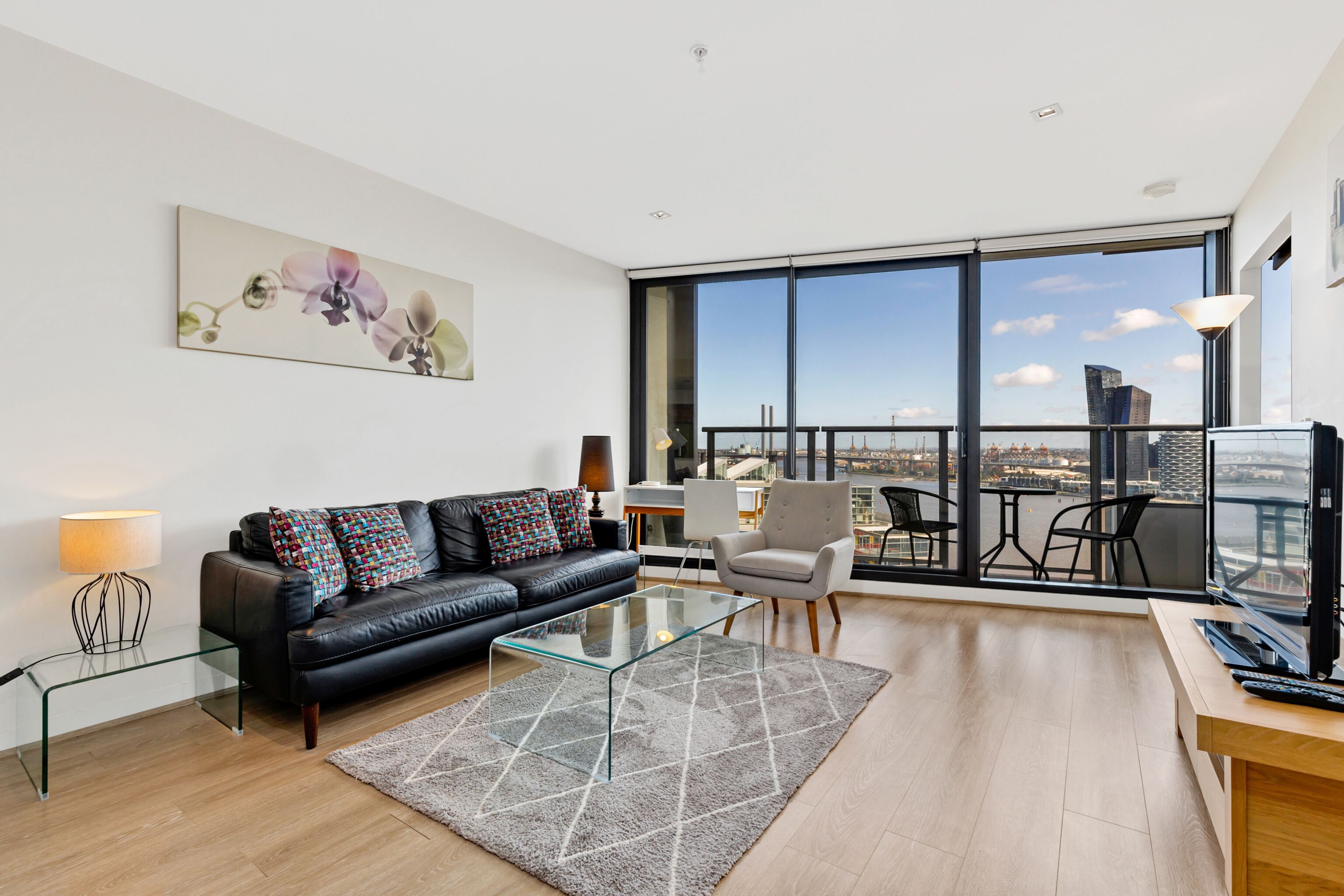 A modern living room with a black leather sofa, a glass coffee table, and a view of the city through large windows.