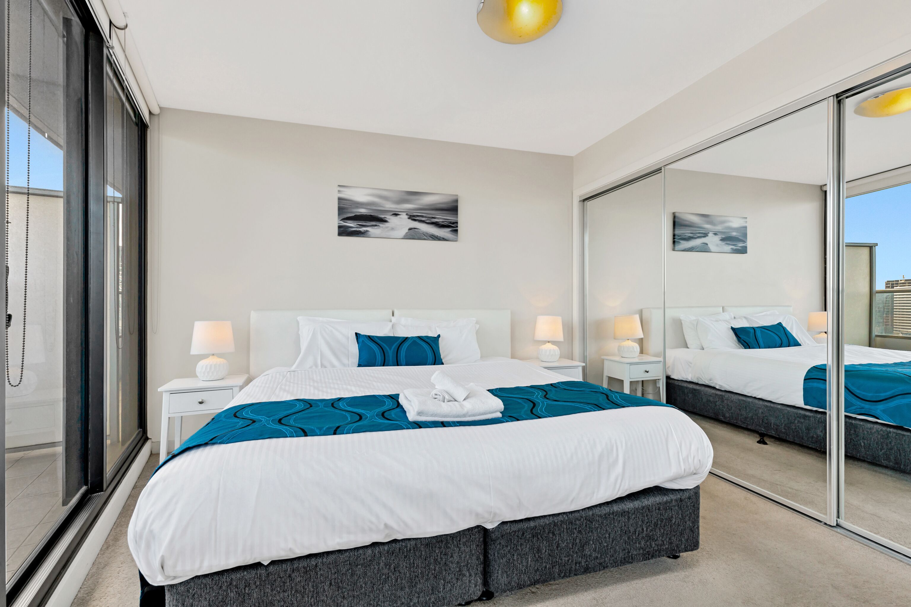 Quarto de hotel moderno com uma cama grande, uma mesinha de cabeceira com abajur, um espelho amplo e vista para a cidade pela janela.