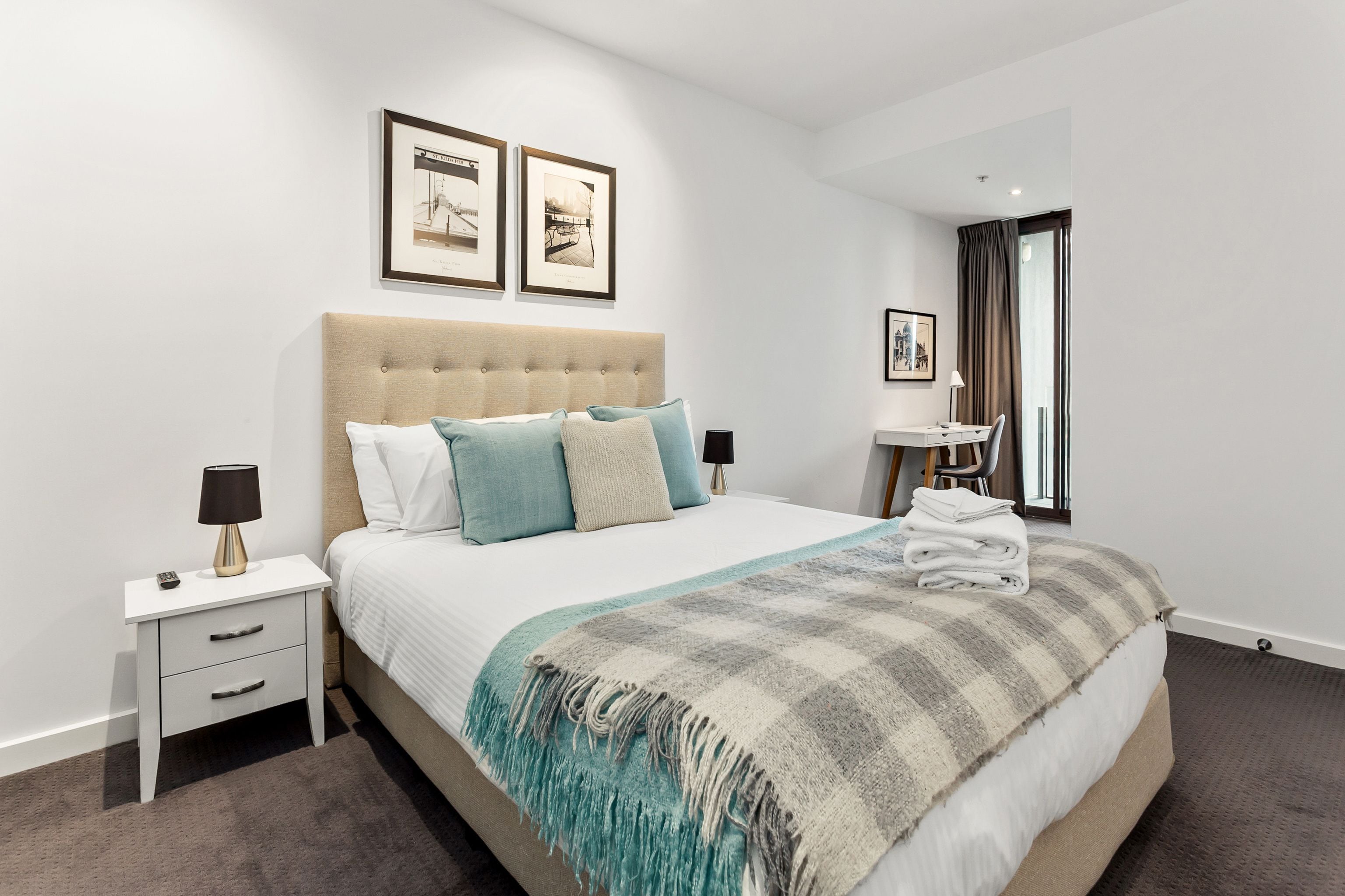 A neatly arranged hotel room with a large bed, a bedside table with a lamp, a small table with a chair, and framed pictures on the wall.