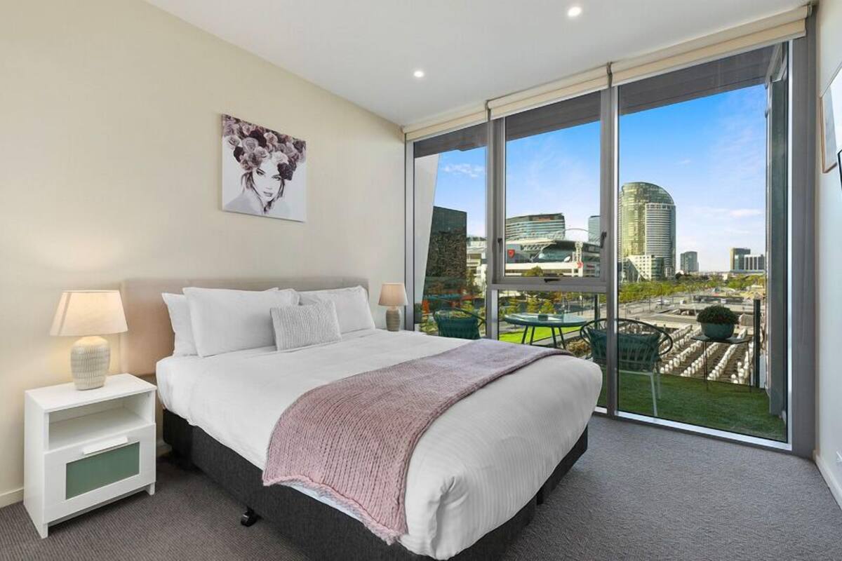A modern hotel room with a large bed, a nightstand, a lamp, and a city view through floor-to-ceiling windows.