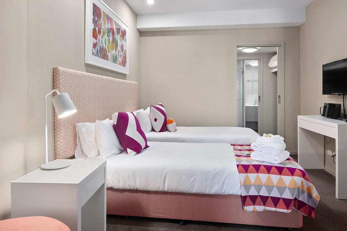 A hotel room with two beds, a desk, a TV, and a bathroom visible through an open door.