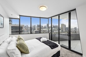 A modern bedroom with a city view, a large bed, and a sliding glass door to an outdoor balcony. - Docklands Private Collection of Apartments - NewQuay (Docklands)