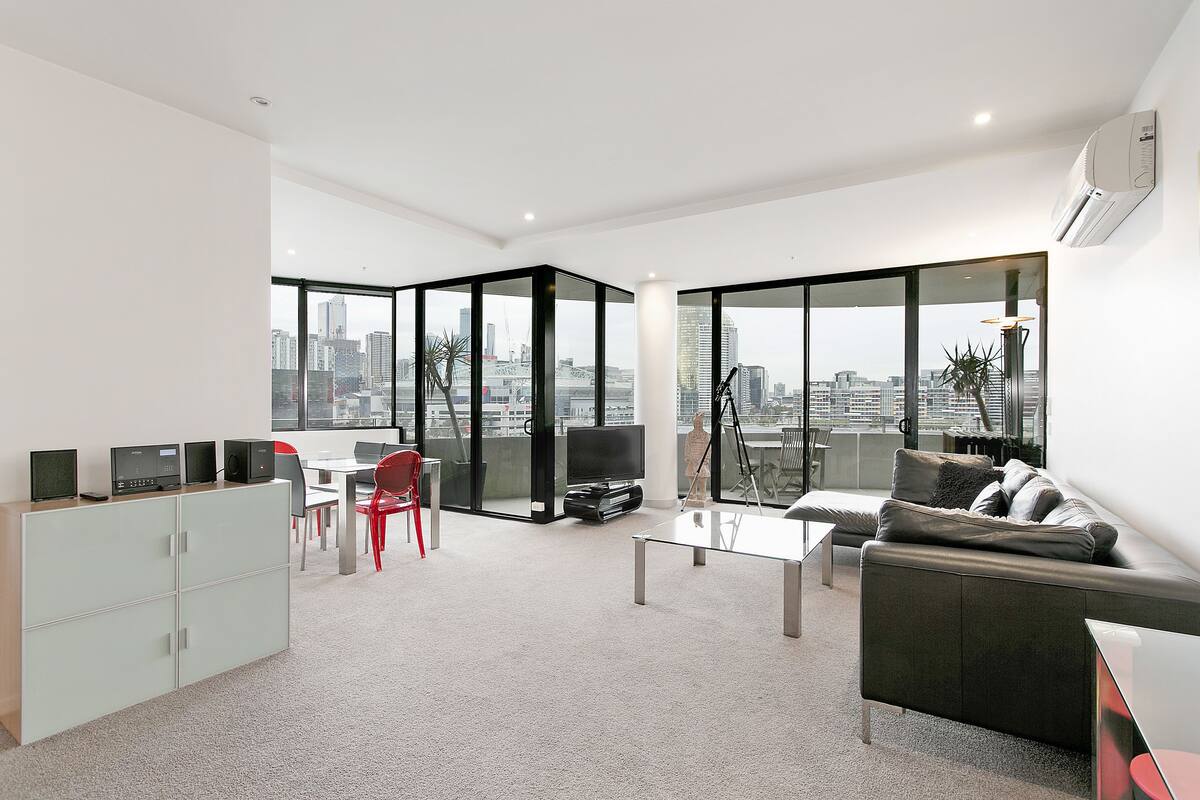 A modern living room with a sofa, a coffee table, and a TV. There is a dining area with a table and chairs. The room has large windows with a city view.