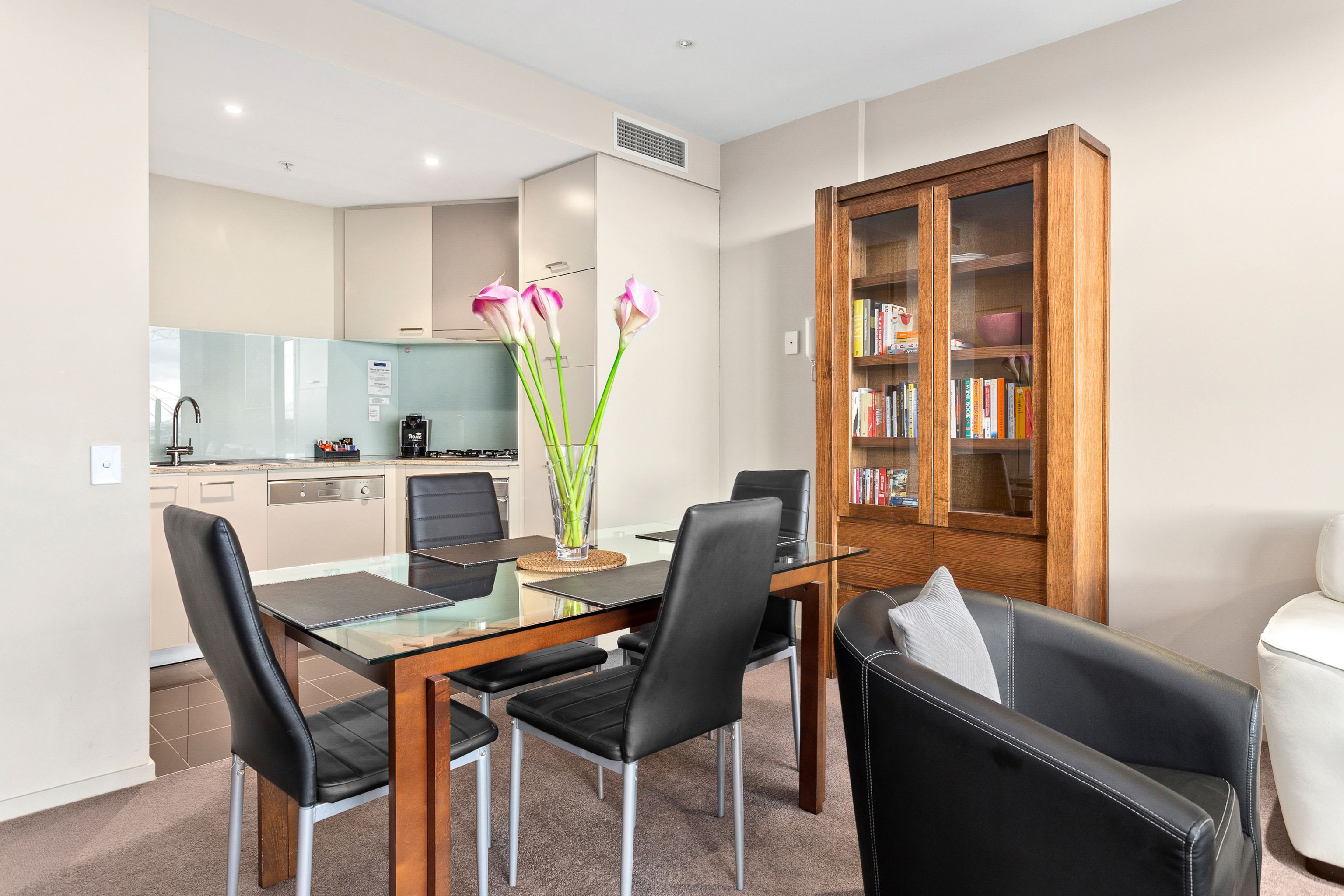 A modern kitchen with a glass-top dining table, black chairs, and a wooden bookshelf.