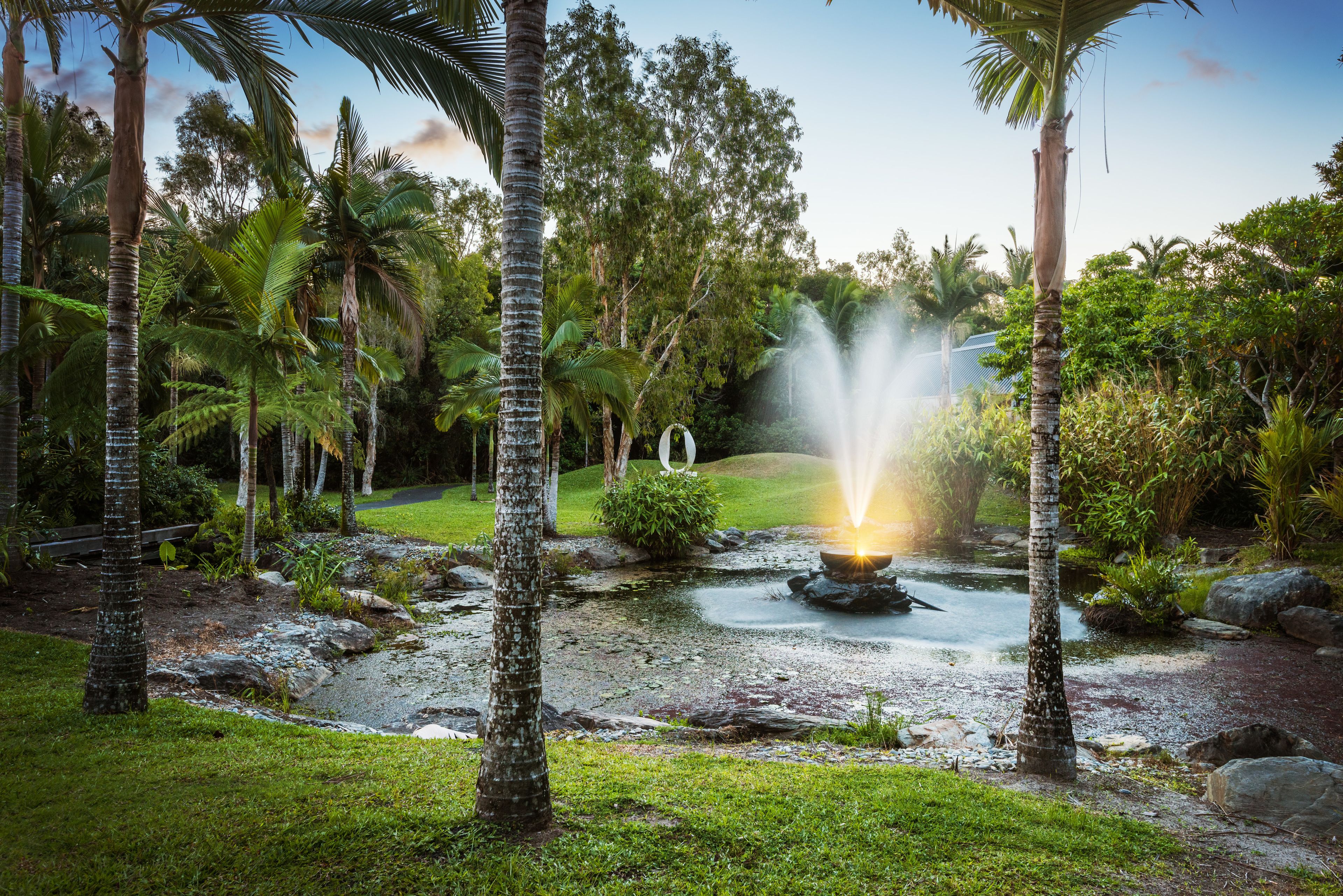 A serene garden with a central water feature, surrounded by lush greenery and palm trees.