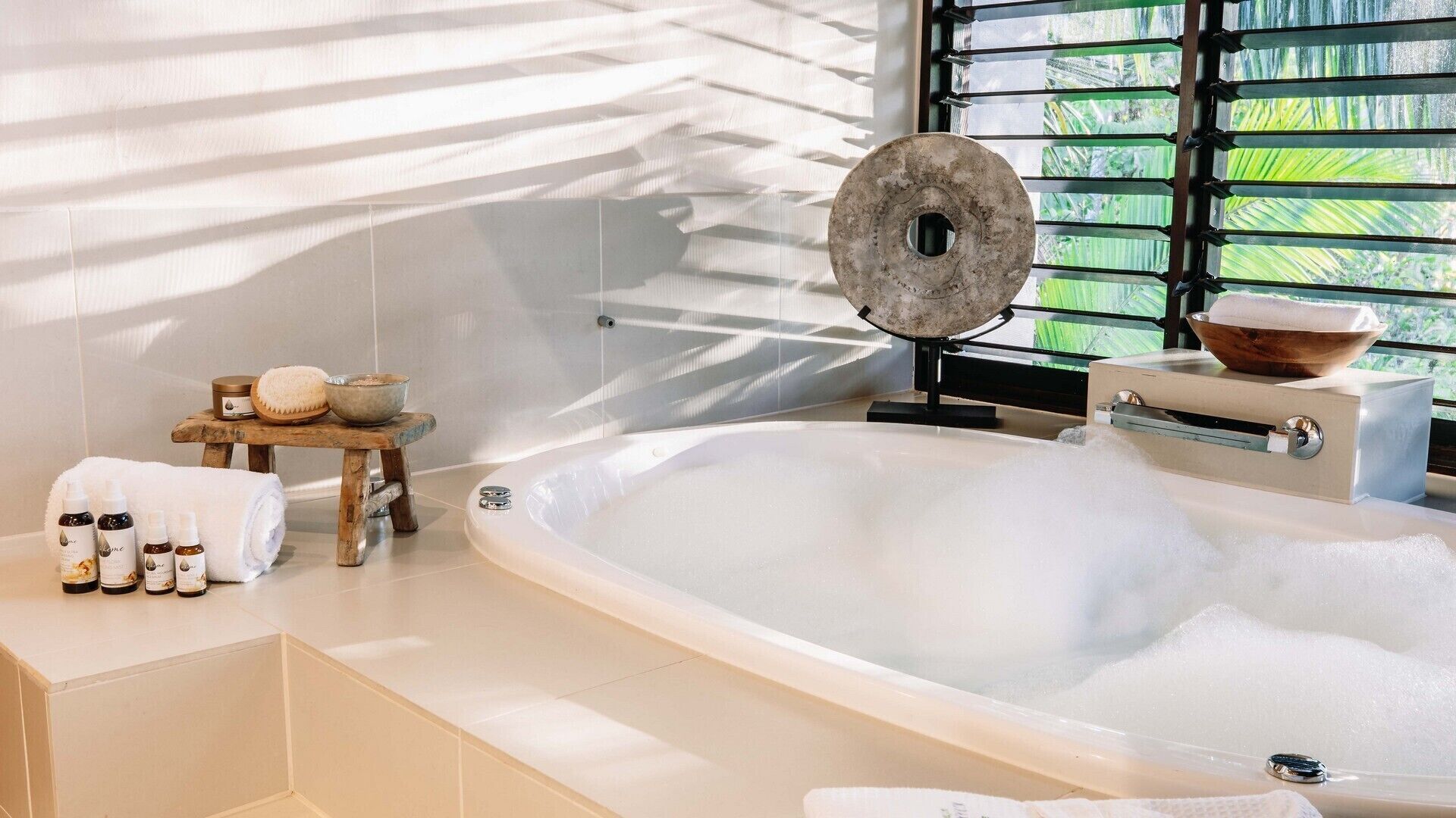 Ein modernes Badezimmer mit einer großen Badewanne voller Blasen, einem hölzernen Hocker mit Toilettenartikeln und einem Fenster mit Grünpflanzen draußen.