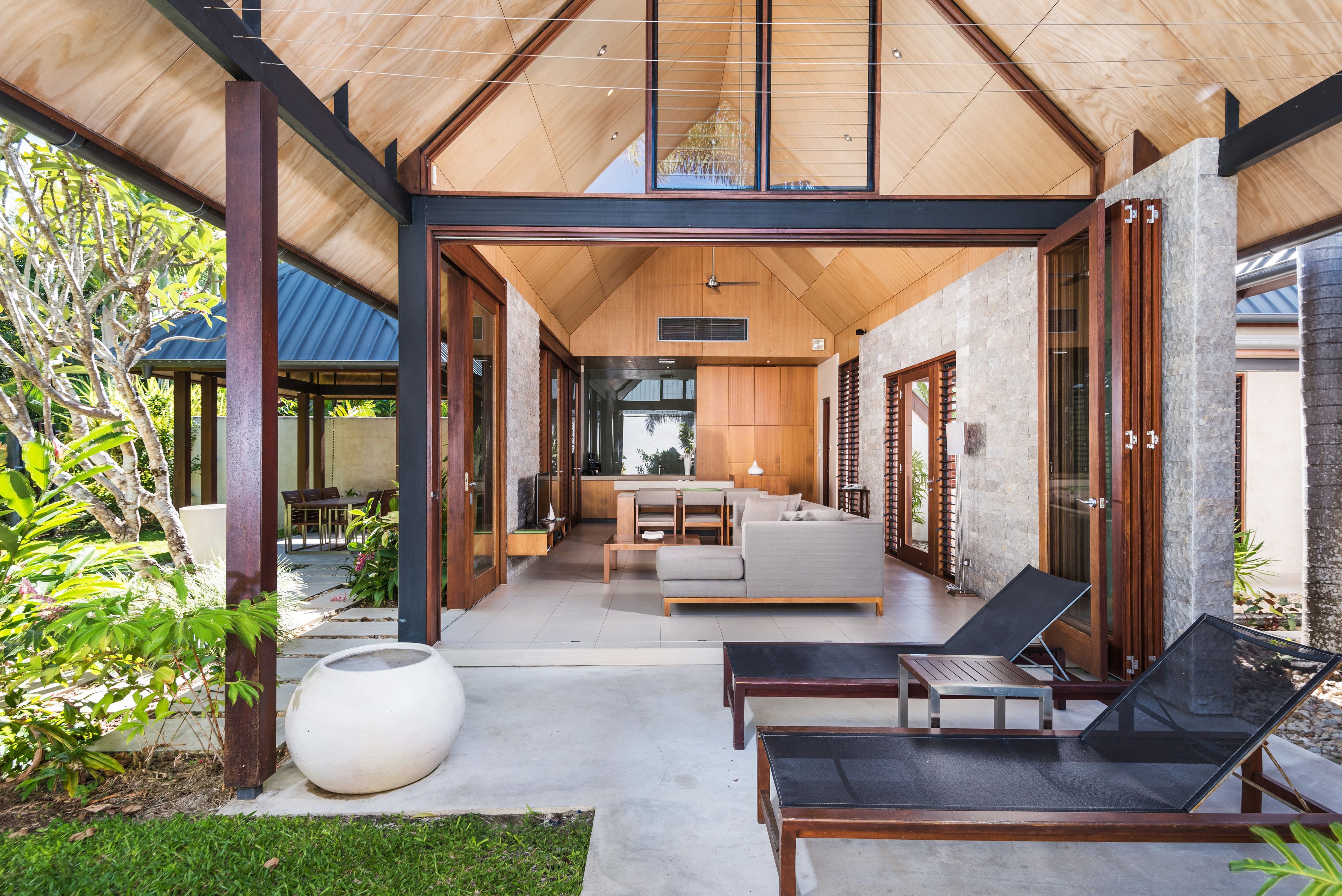 A modern outdoor living area with a wooden ceiling, a large white planter, and a seating arrangement including a sofa and two lounge chairs.