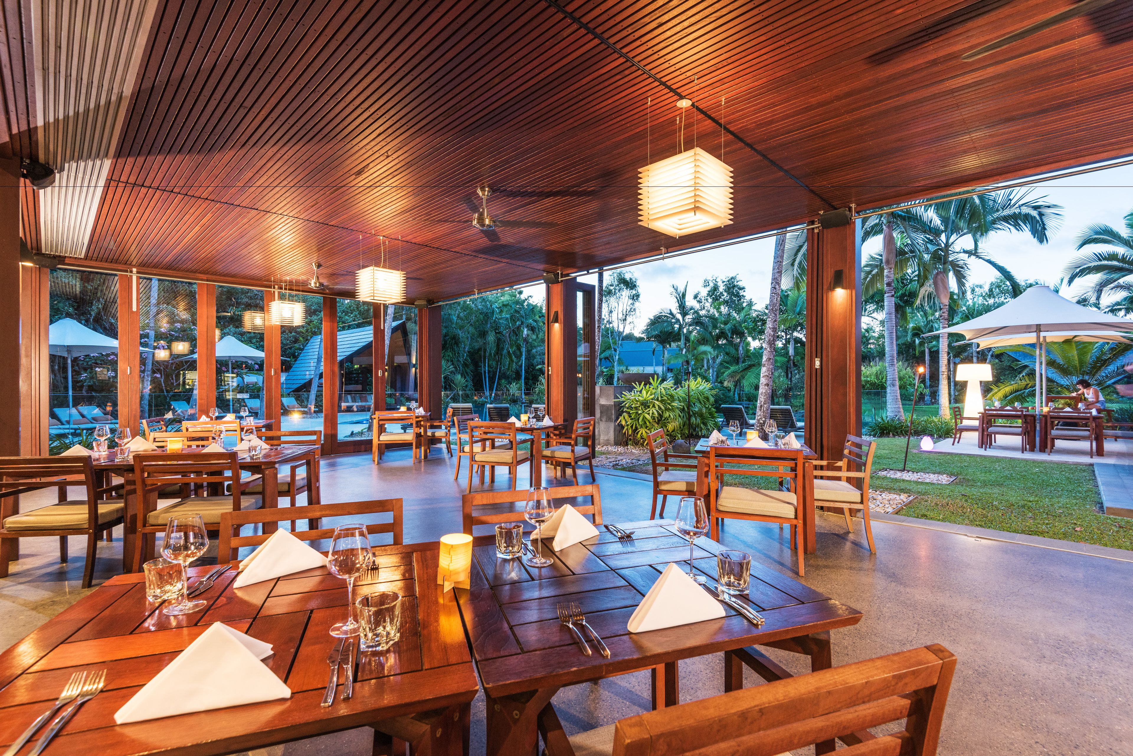 Un área de comedor al aire libre con mesas y sillas de madera, amplias ventanas y vistas a palmeras y follaje.