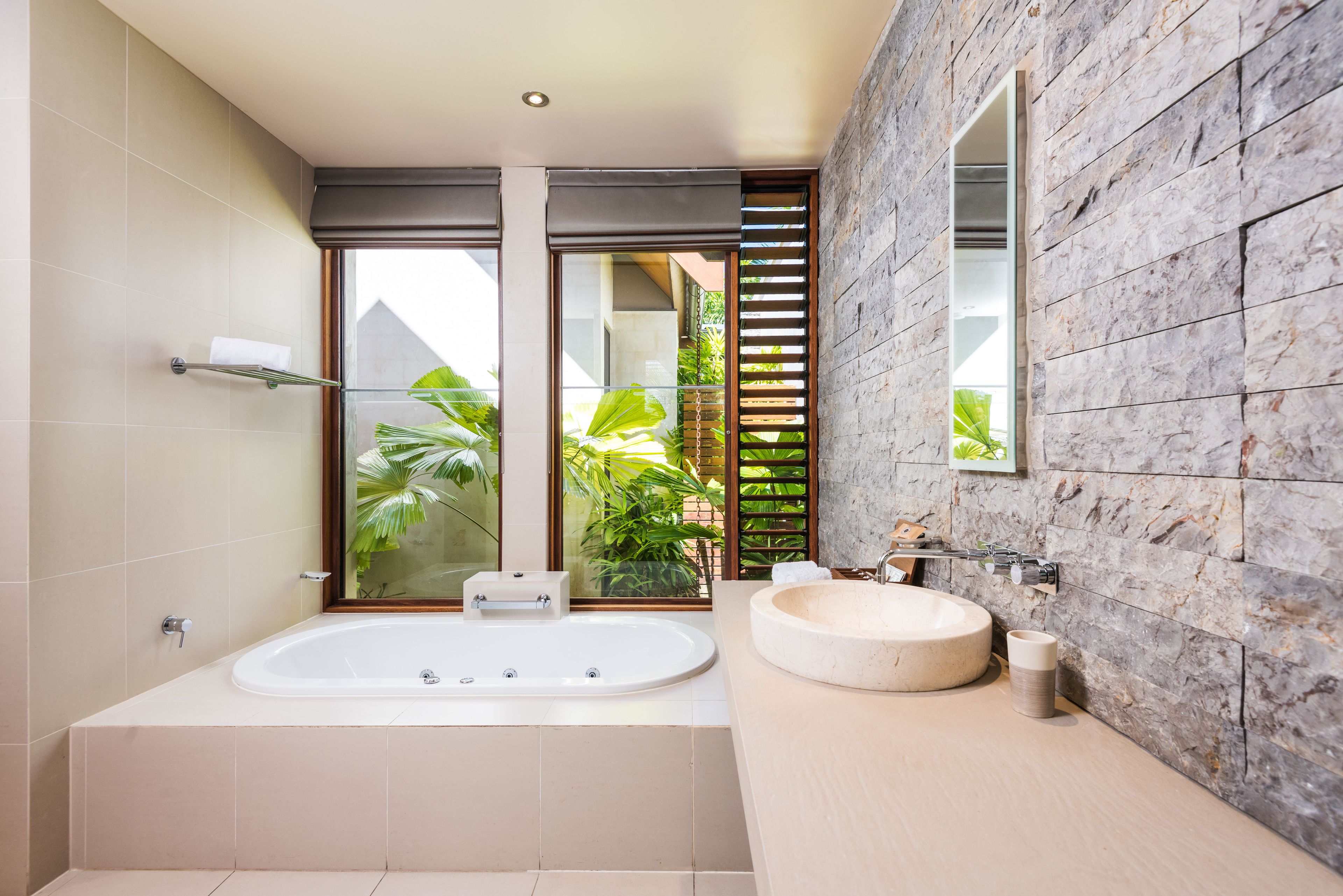 A modern bathroom with a large bathtub, a round sink, and a window with a view of greenery.