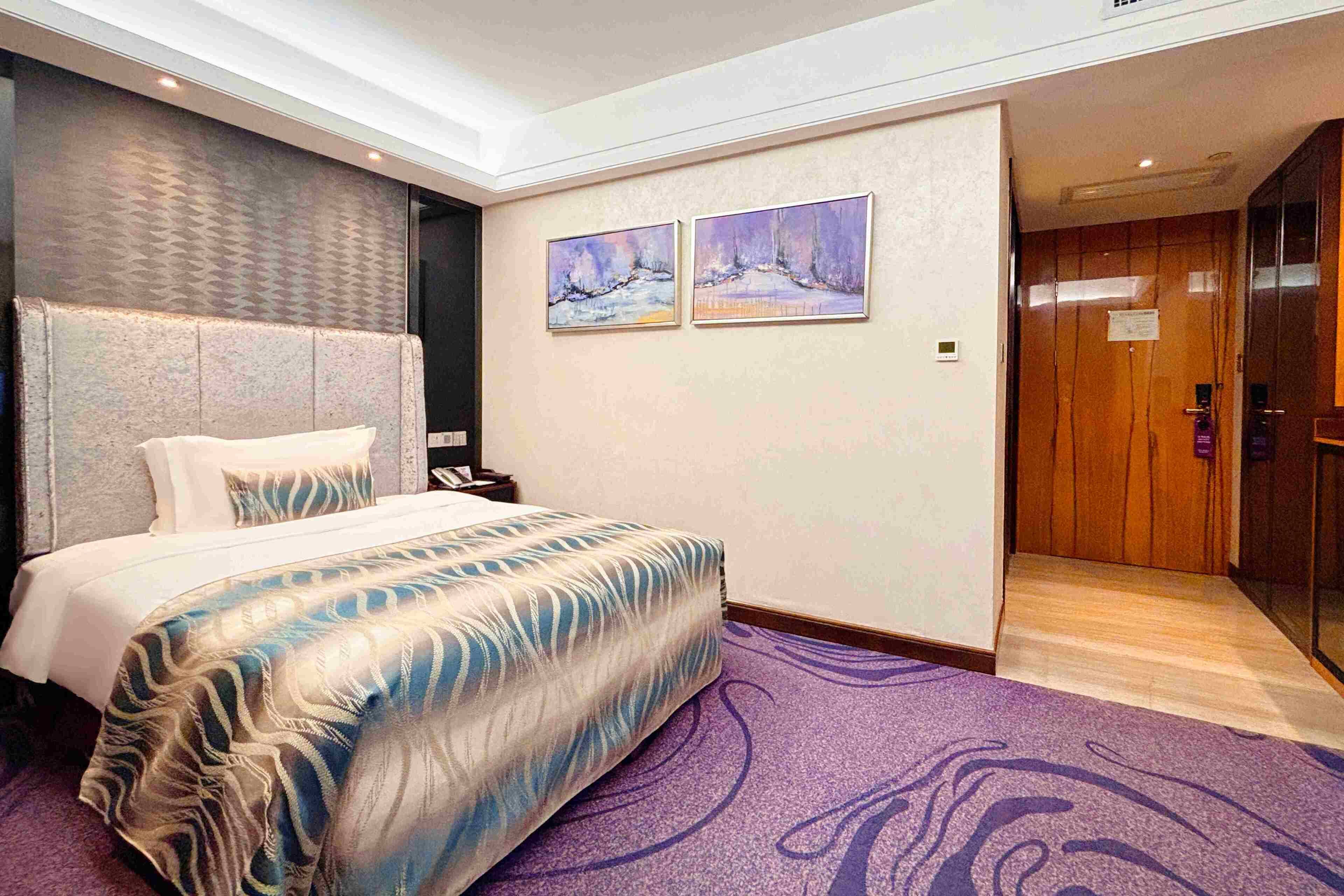 A hotel room with a large bed, a headboard, a purple carpet, and two paintings on the wall.