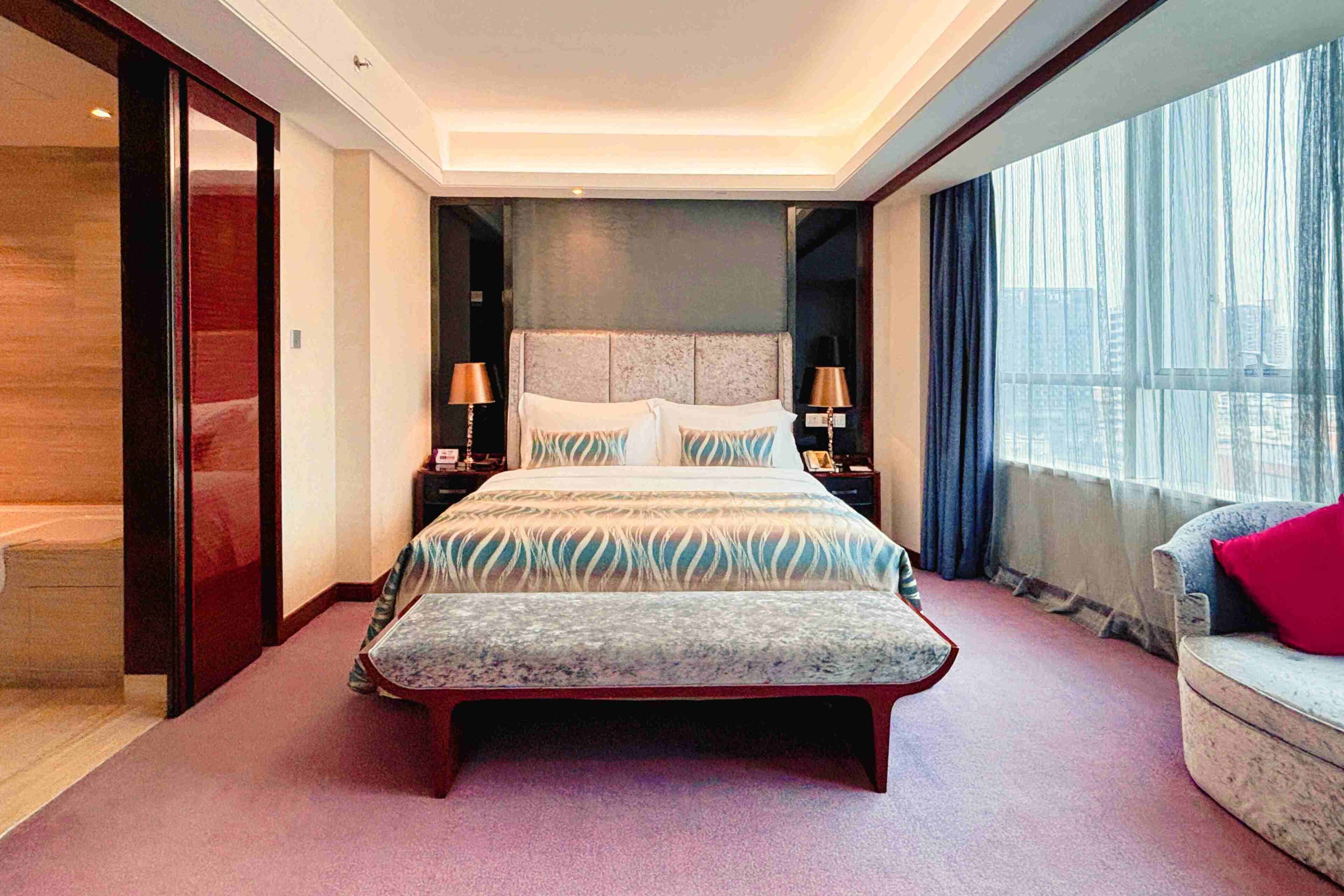 A modern hotel room with a large bed, bedside lamps, a sofa, and a view of the city through the window.