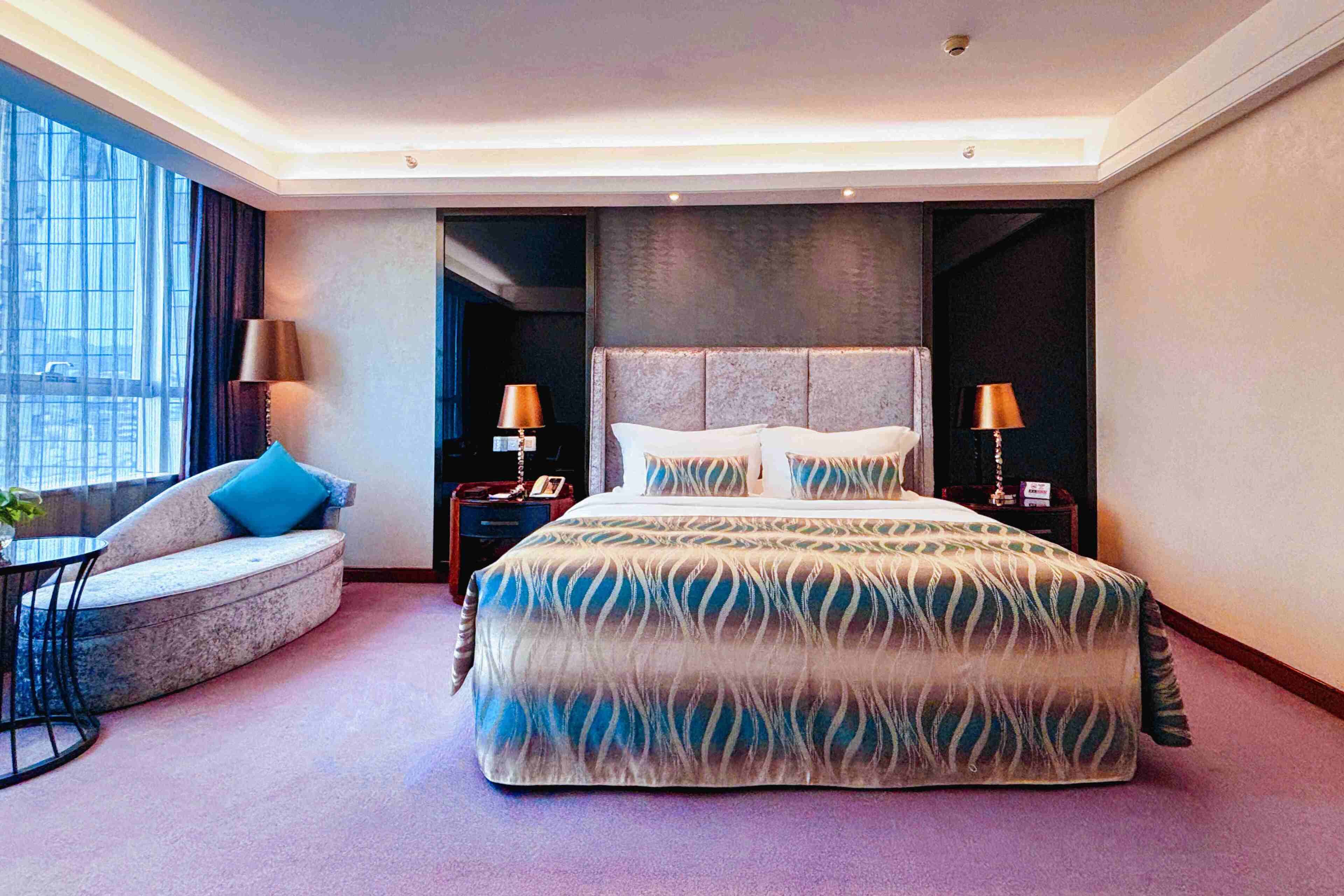 A modern hotel room with a large bed, a sofa, two nightstands, and a window with curtains.