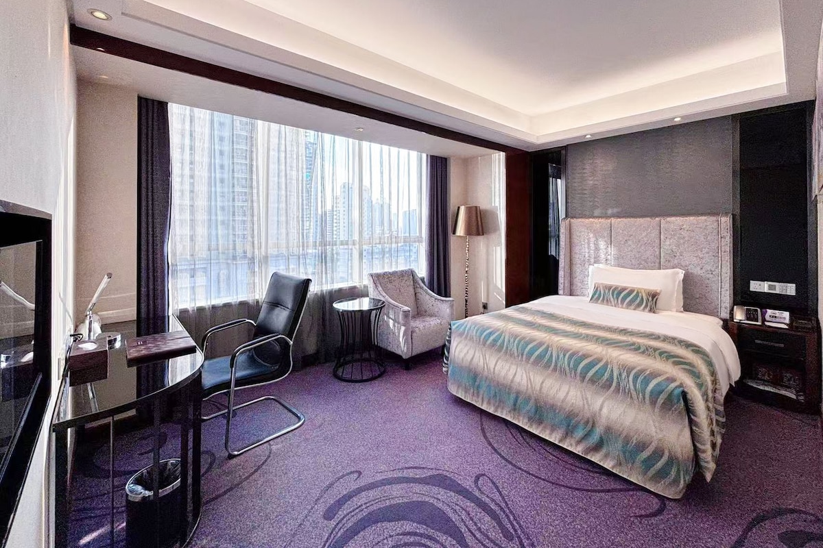 A hotel room with a large bed, a desk, a chair, and a view of the city.