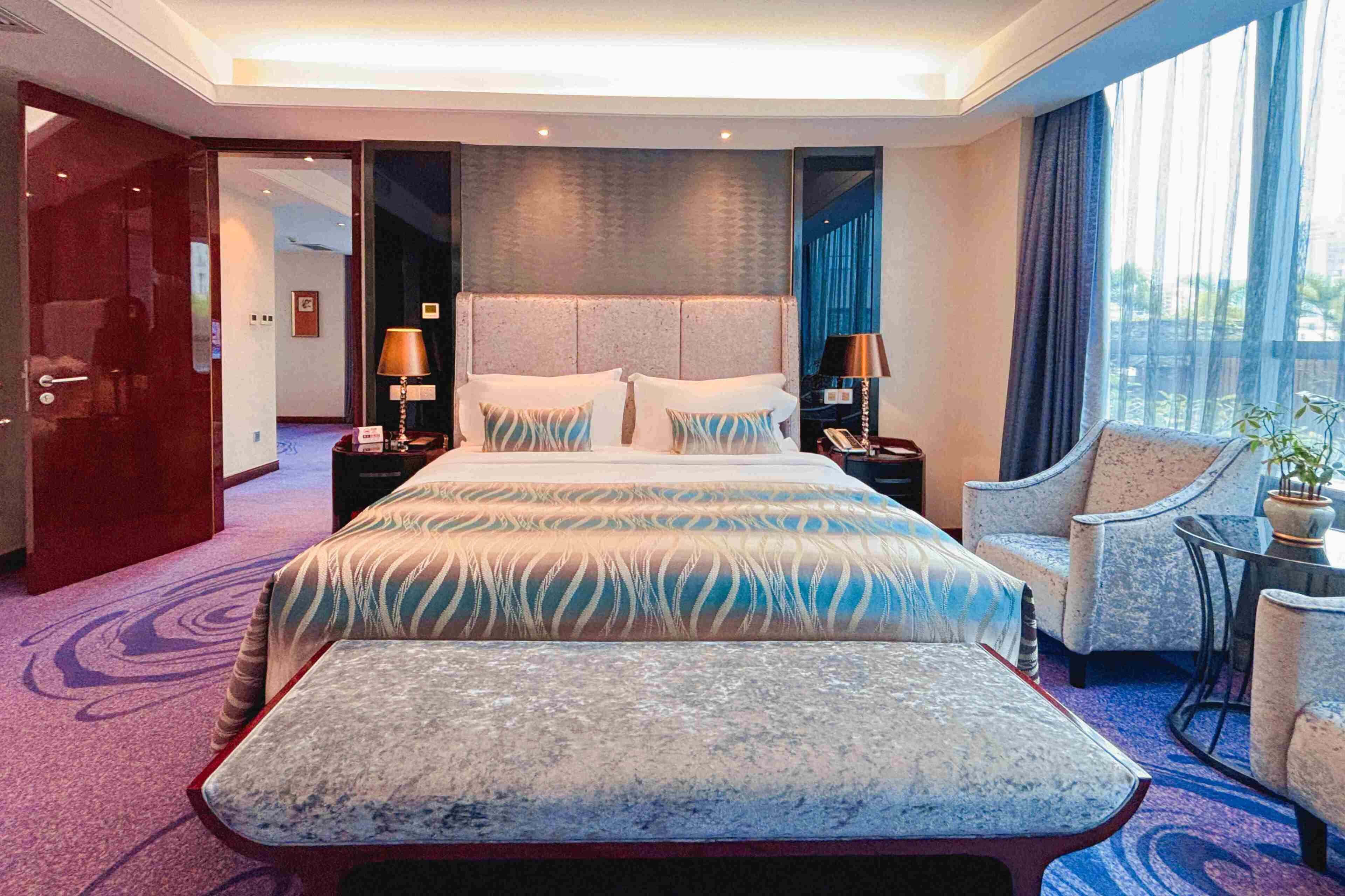 A hotel room with a large bed, two bedside tables with lamps, a chair, a small table, and a view of the outdoors through a window.