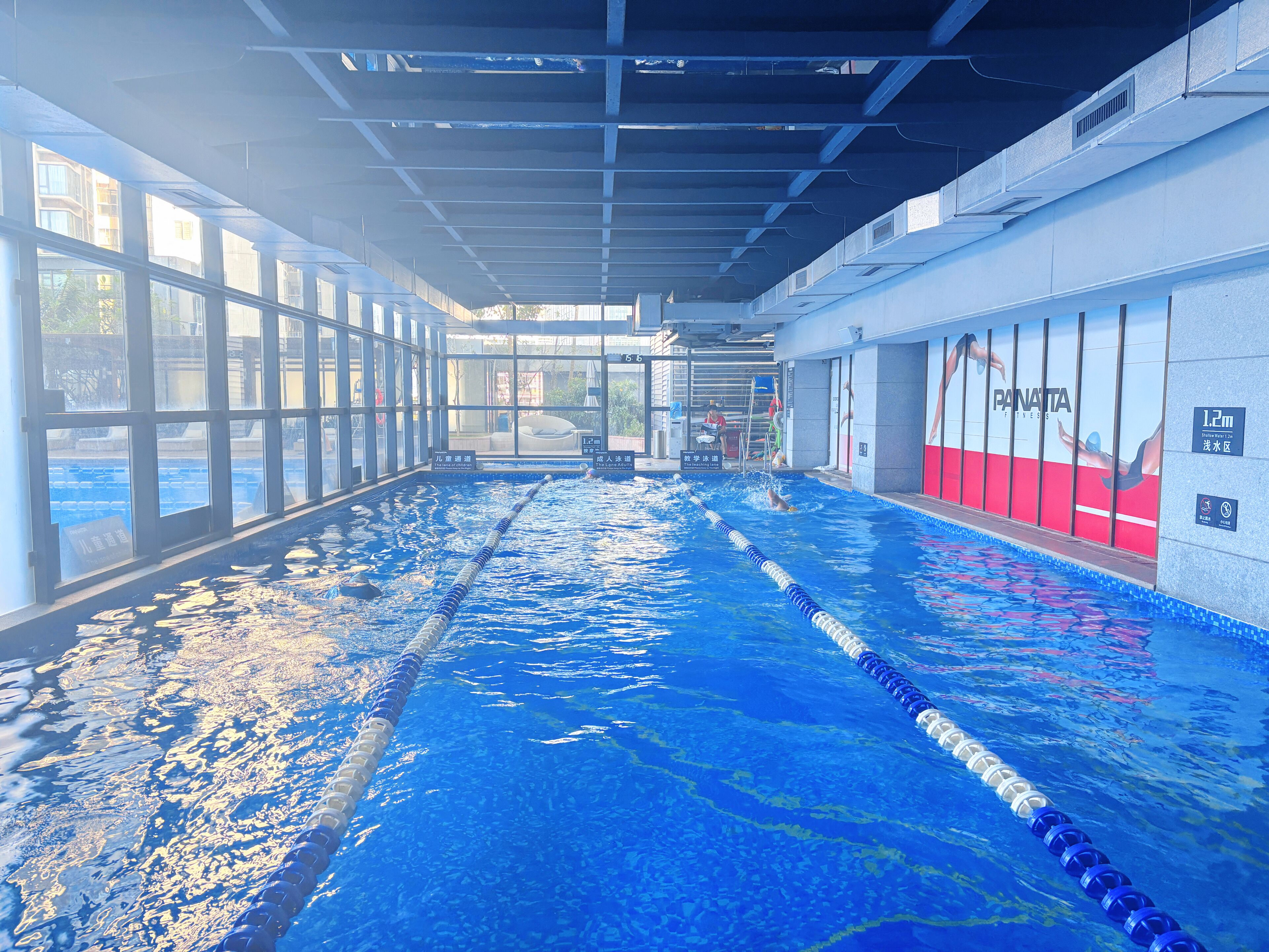 Sisätiloissa sijaitseva uima-allas, jossa on selkeästi merkityt uimakaistat, suuret ikkunat sekä punavalkoinen seinä, jossa on näkyvillä mainos tekstillä ”Panatta Fitness”.