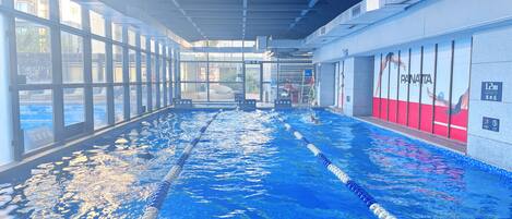 Sisätiloissa sijaitseva uima-allas, jossa on selkeästi merkityt uimakaistat, suuret ikkunat sekä punavalkoinen seinä, jossa on näkyvillä mainos tekstillä ”Panatta Fitness”.