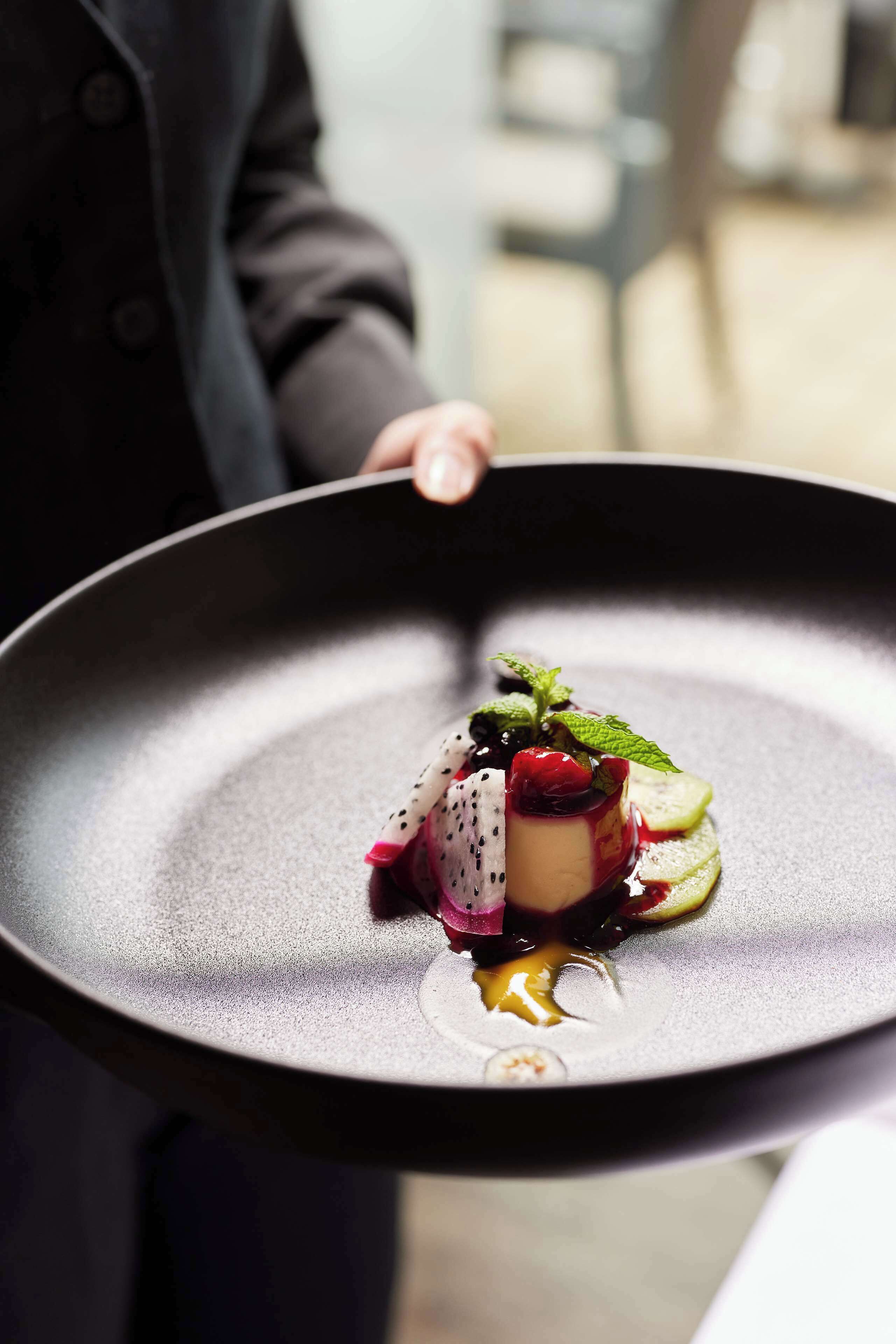 Une personne tient une assiette noire sur laquelle se trouve un dessert composé d’une garniture de fruits, de feuilles de menthe et d’une sauce.