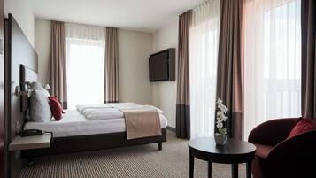 Ein Hotelzimmer mit einem Bett, einem Schreibtisch mit Telefon, einem an der Wand montierten Fernseher, einem kleinen Tisch mit Pflanze und einem Stuhl.