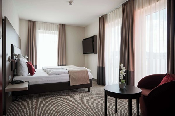 A hotel room with a bed, a desk with a telephone, a TV mounted on the wall, a small table with a plant, and a chair. - bigBOX ALLGÄU Hotel, WorldHotels Distinctive (Kempten)