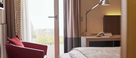 Une chambre d’hôtel avec un lit, une chaise rouge, un bureau avec une lampe et une vue sur l’extérieur, visible à travers une porte ouverte.