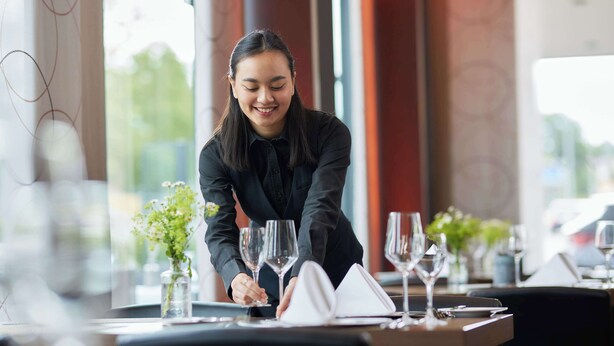 Una persona preparando una mesa con copas de vino y una carta en un restaurante con amplios ventanales y una decoración moderna.