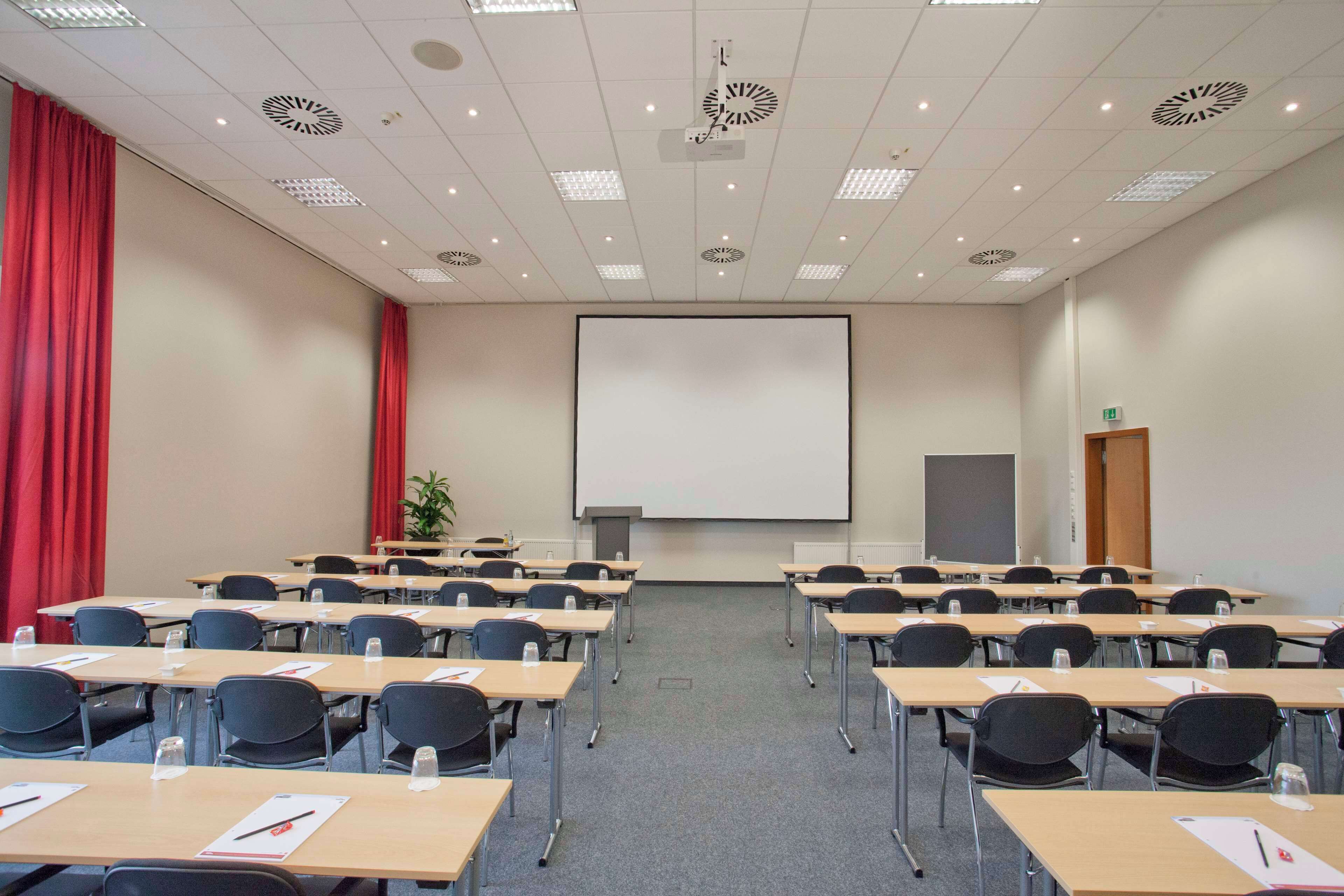 Uma sala de conferências com mesas e cadeiras dispostas em fileiras, uma tela de projeção grande e uma cortina vermelha em um dos lados.