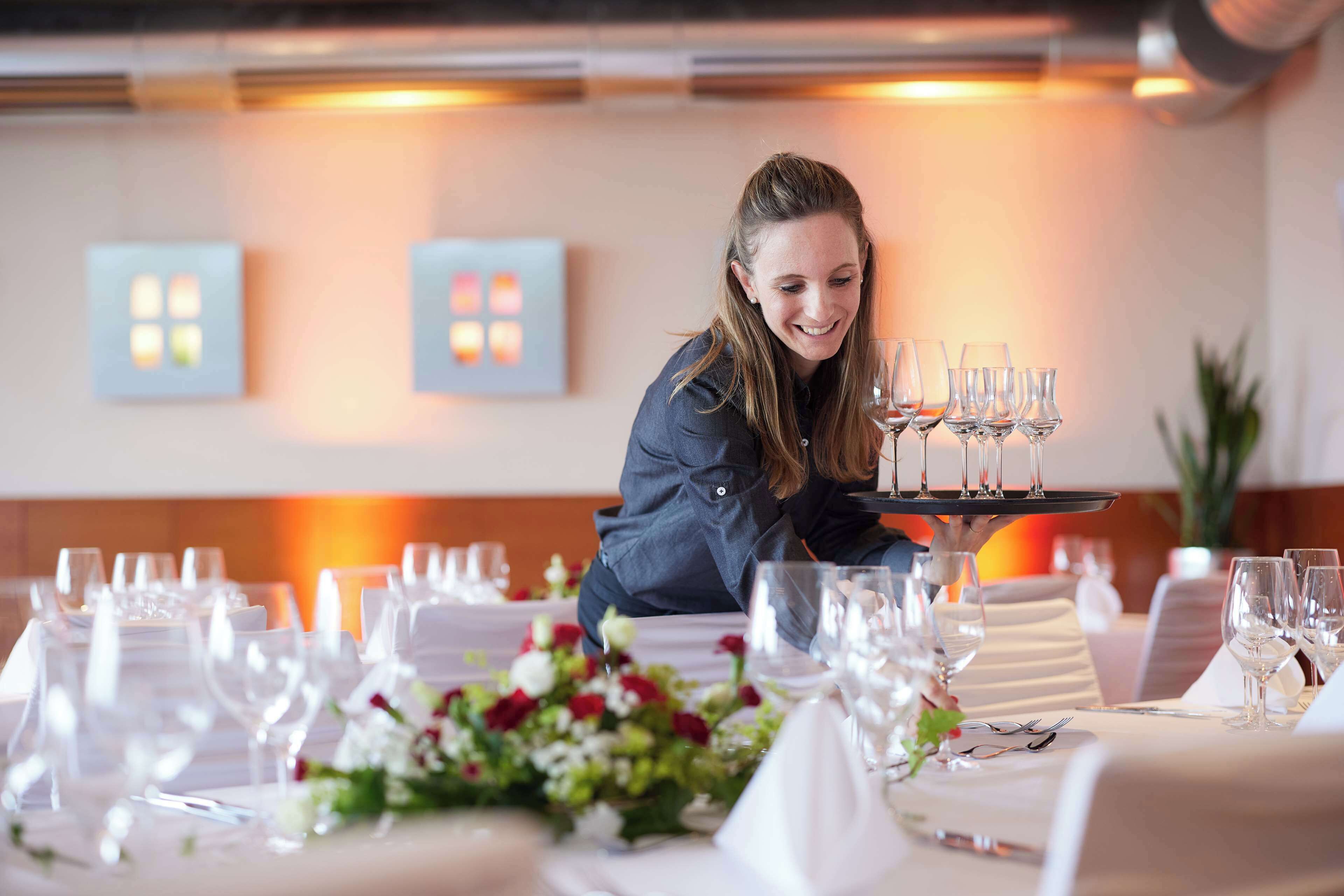 A well-set dining table with multiple wine glasses and a tray, a floral centerpiece, and a modern interior with wall-mounted light fixtures.